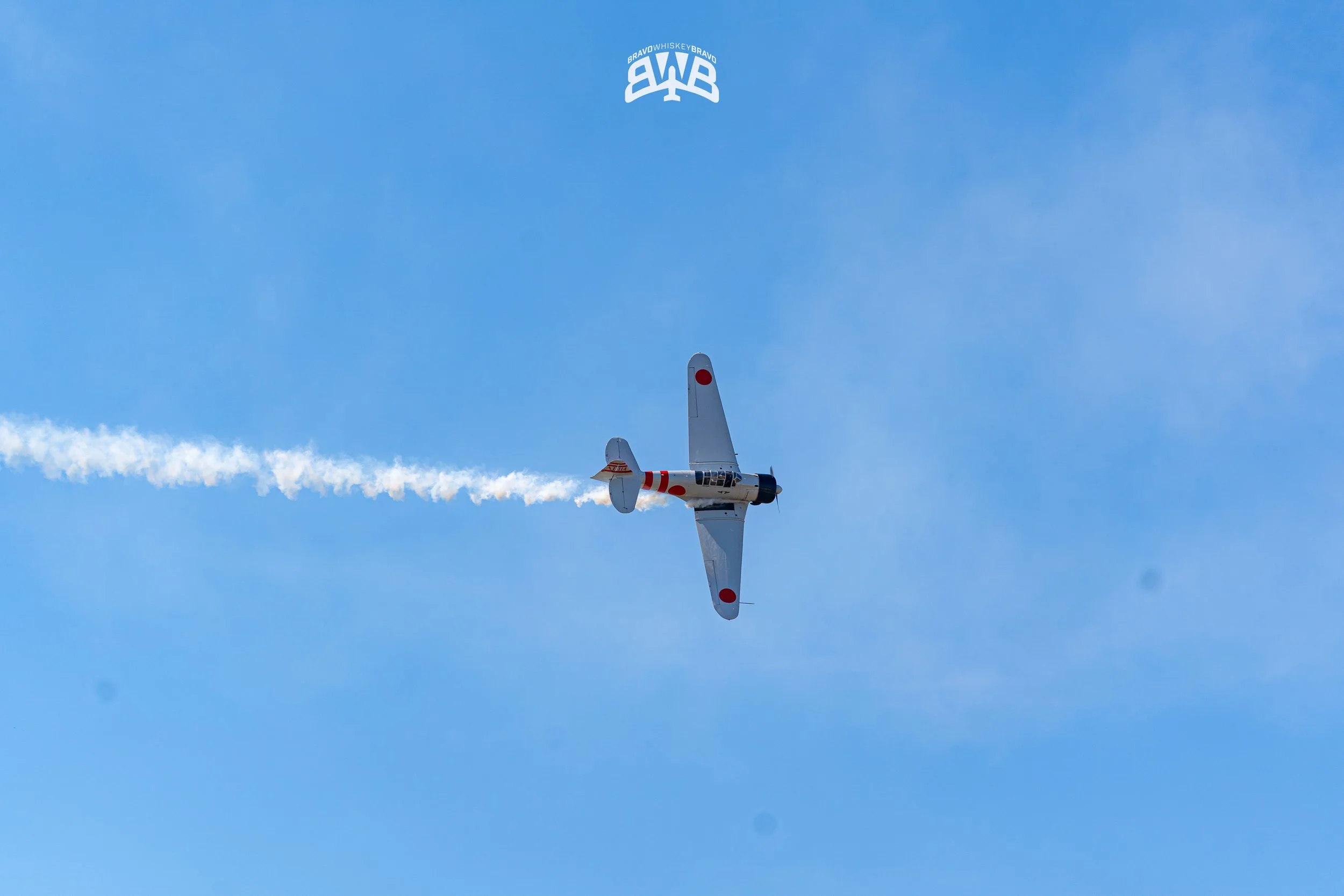 A vintage propeller airplane with red and white markings flying through a blue sky, leaving a white smoke trail behind.
