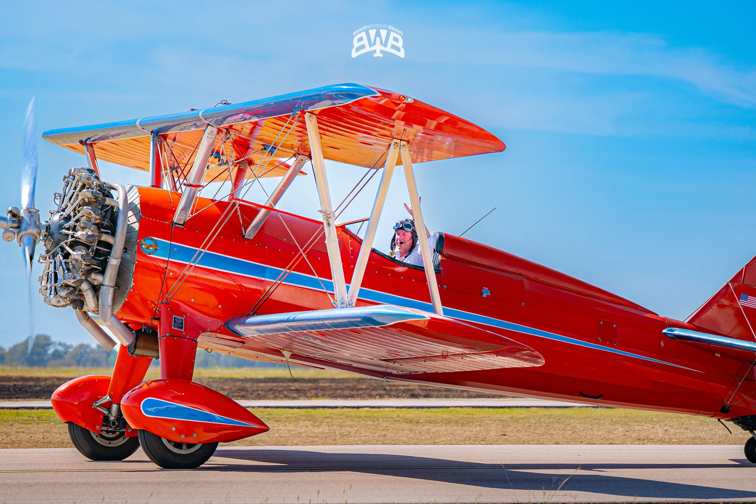 A bright red vintage biplane with a smiling pilot, positioned on a runway with blue sky in the background.