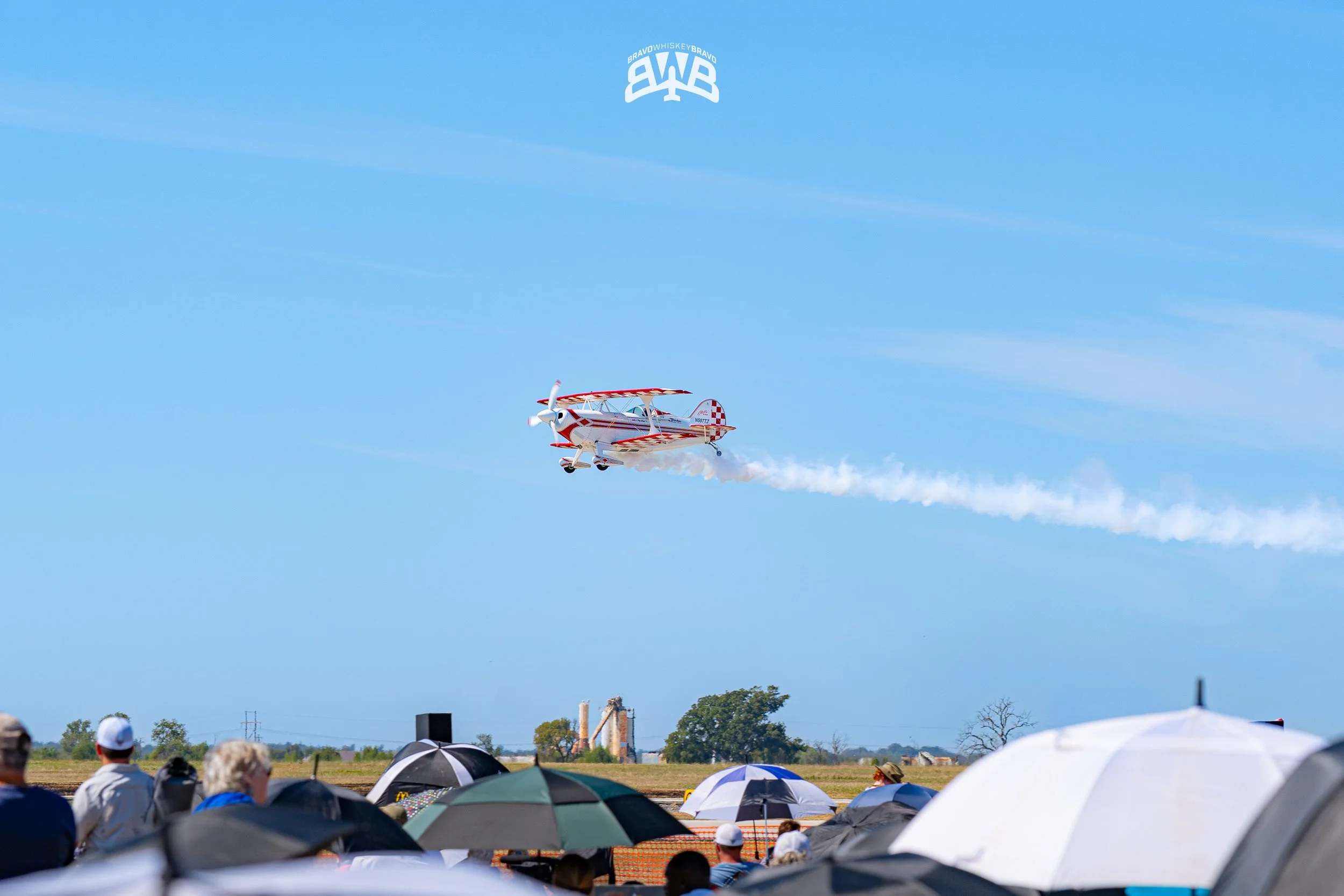 An airplane performing an aerobatic stunt in the sky with smoke trail behind it, spectators with umbrellas watching from the ground, and a clear blue sky overhead.