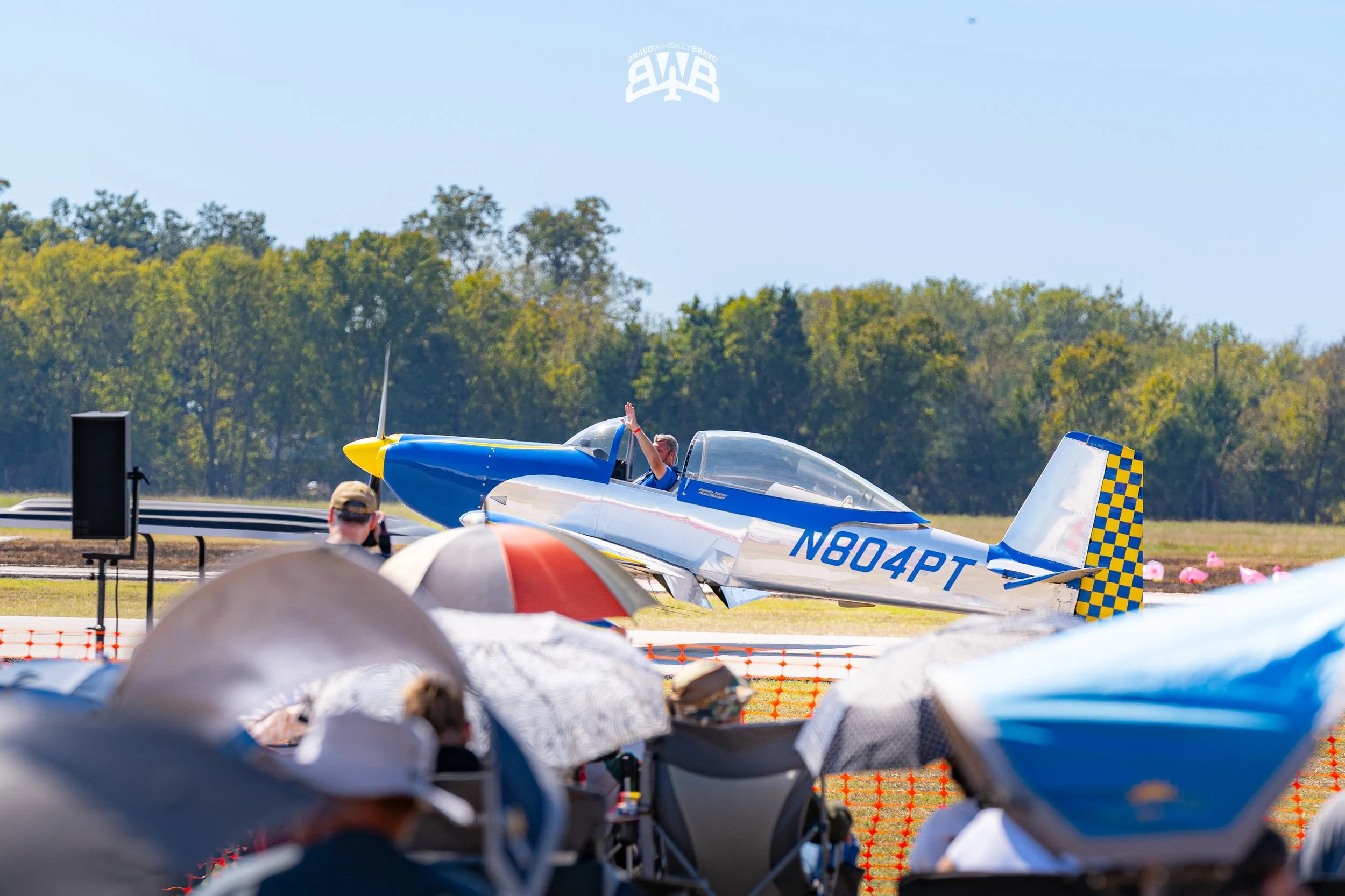 A small aerobatic airplane with blue and white colors and checkered tail, N804PT, is on the ground with a pilot waving to the crowd. Spectators sit under umbrellas watching the event on a sunny day with trees in the background.