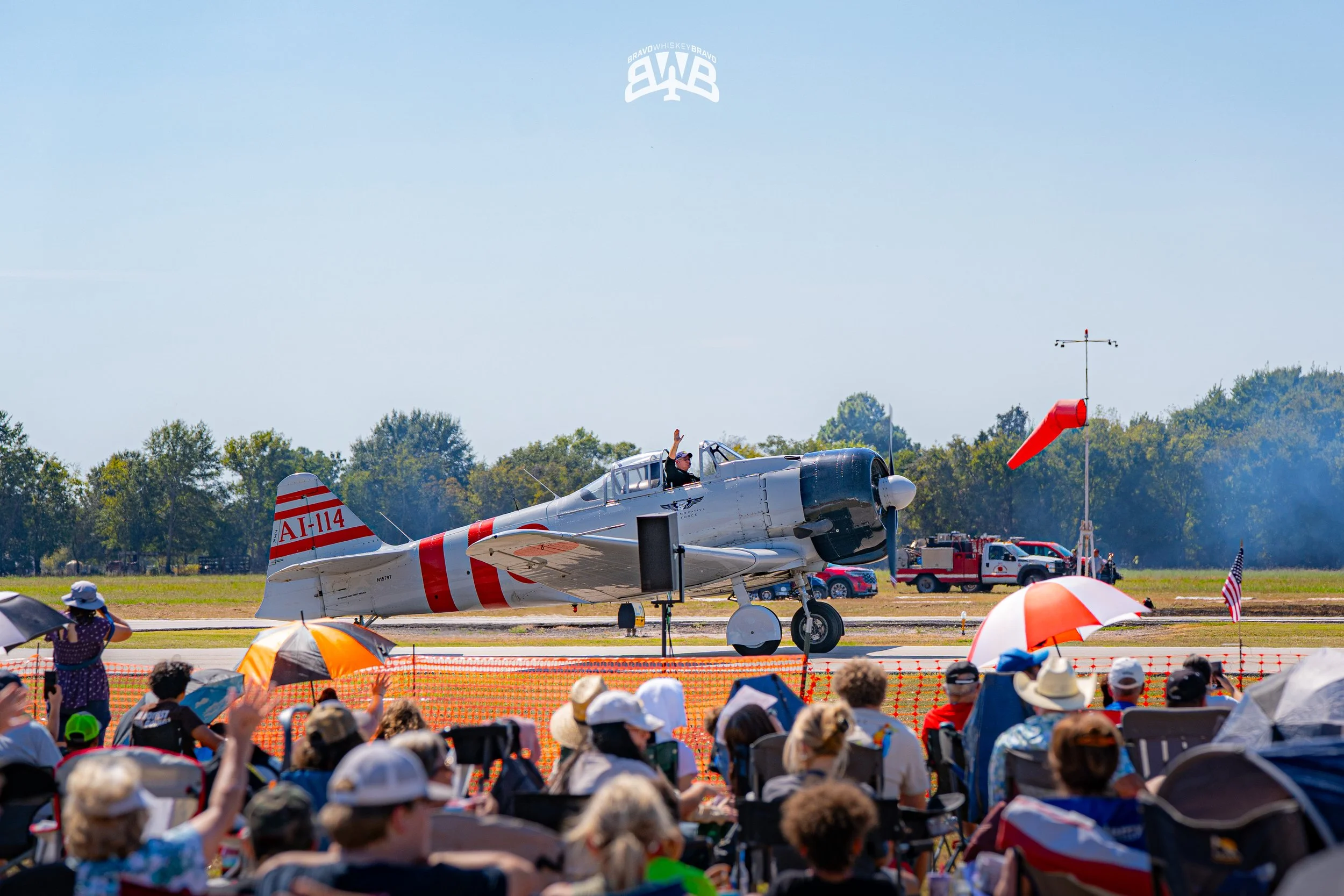 An airshow with a vintage aircraft on the runway and a crowd of spectators in front, some holding umbrellas, with the pilot waving from the open cockpit, and a windsock on the right side of the image.