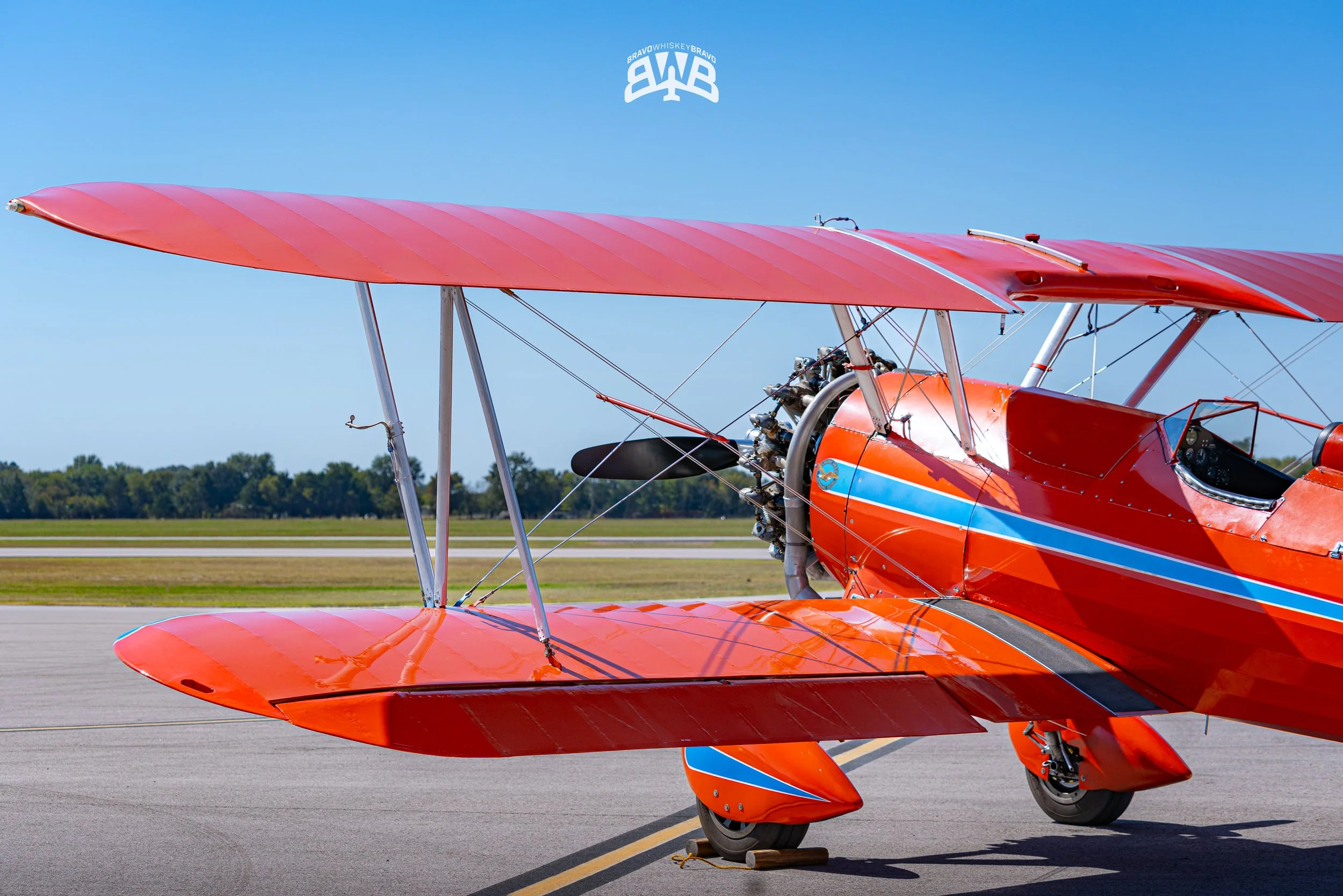 Red biplane parked on an airfield with blue sky and distant trees in the background.