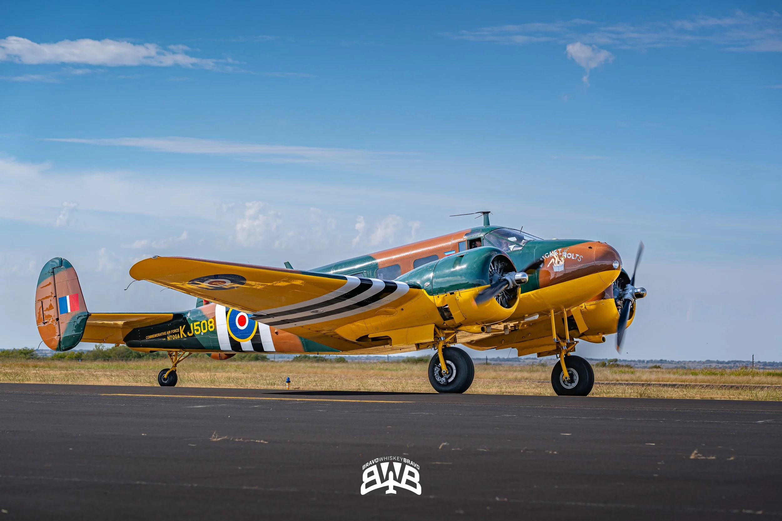 A vintage WWII-era fighter plane painted in camouflage colors with yellow accents, parked on a runway against a blue sky with some clouds.