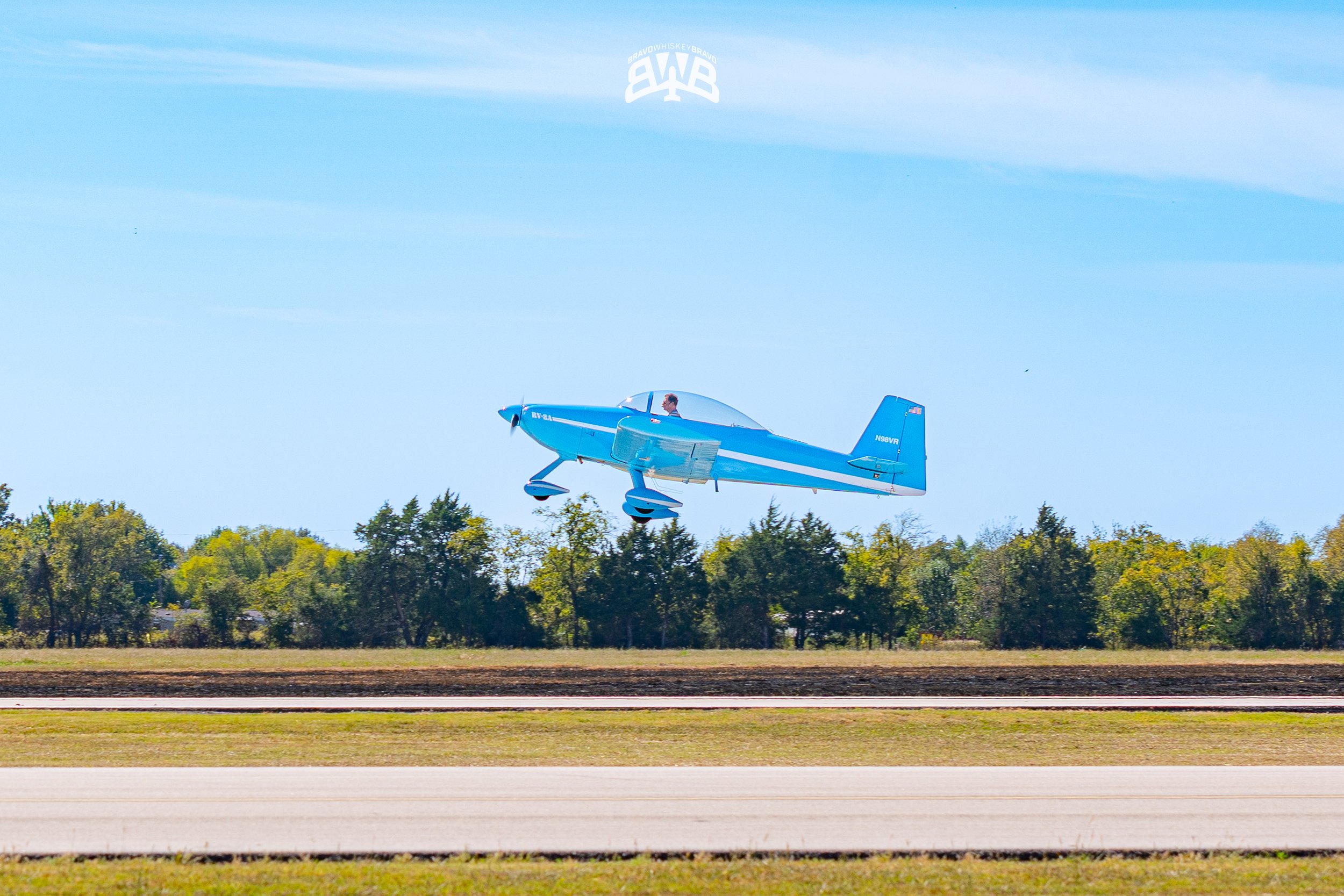 A small blue airplane flying low over a grassy field with trees in the background