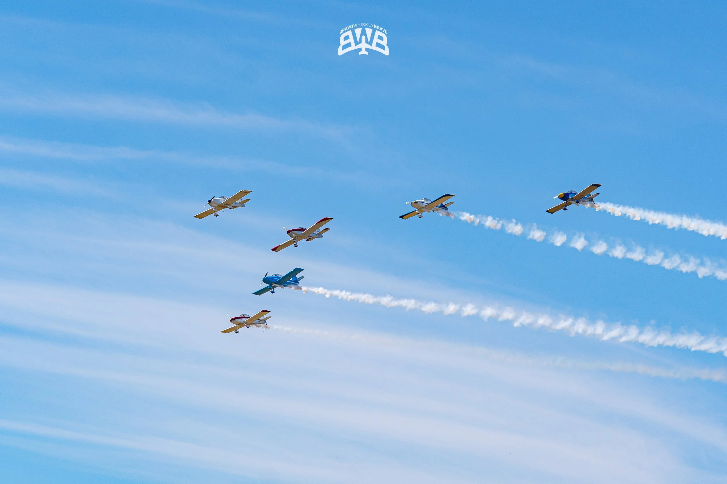 Six small airplanes flying in formation in a clear blue sky, leaving white smoke trails behind them.