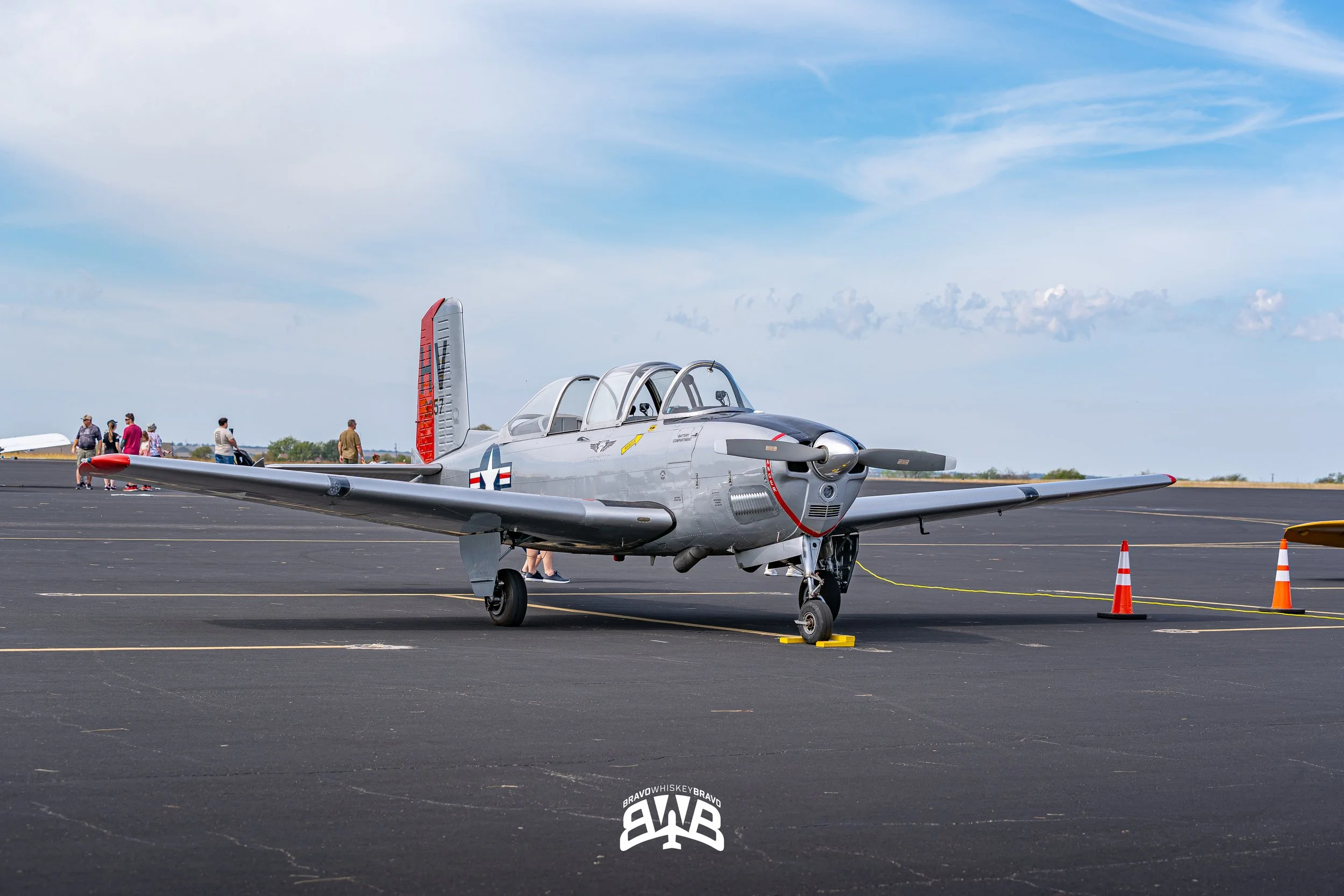 A silver vintage military aircraft parked on an airport tarmac with a few people walking in the background and orange traffic cones nearby.