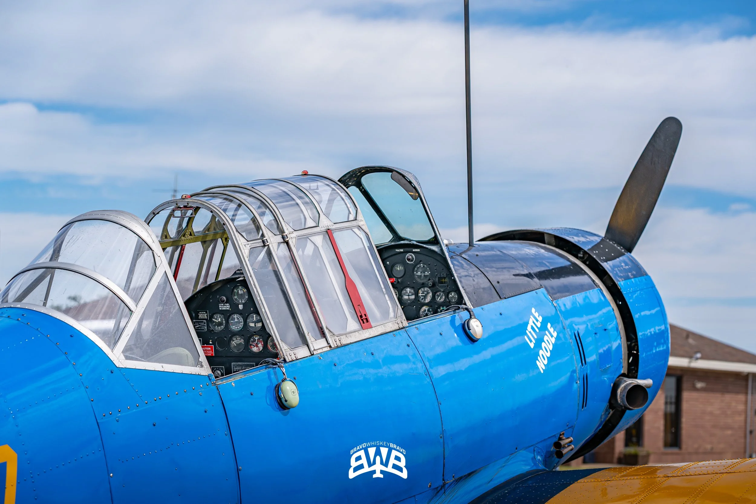 Close-up of the cockpit and nose of a vintage blue military aircraft with a bubble canopy and black propeller, against a partly cloudy sky and residential buildings in the background.