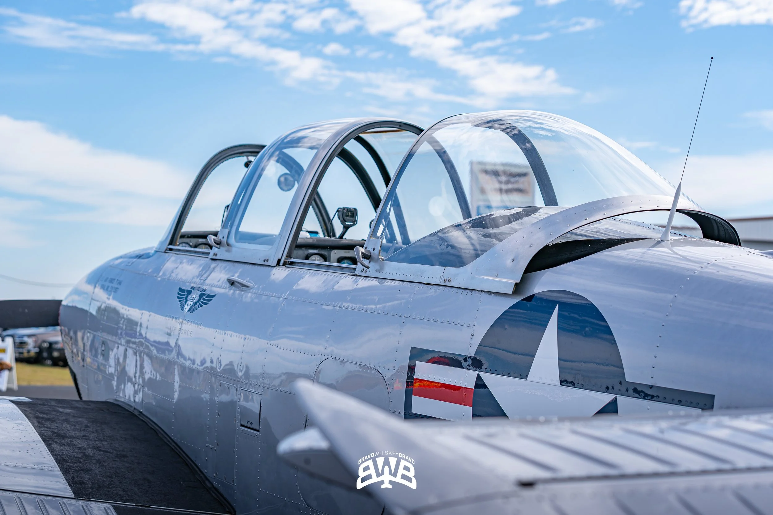 Close-up of a vintage military aircraft with a silver body and top canopy, parked outdoors against a blue sky with clouds.