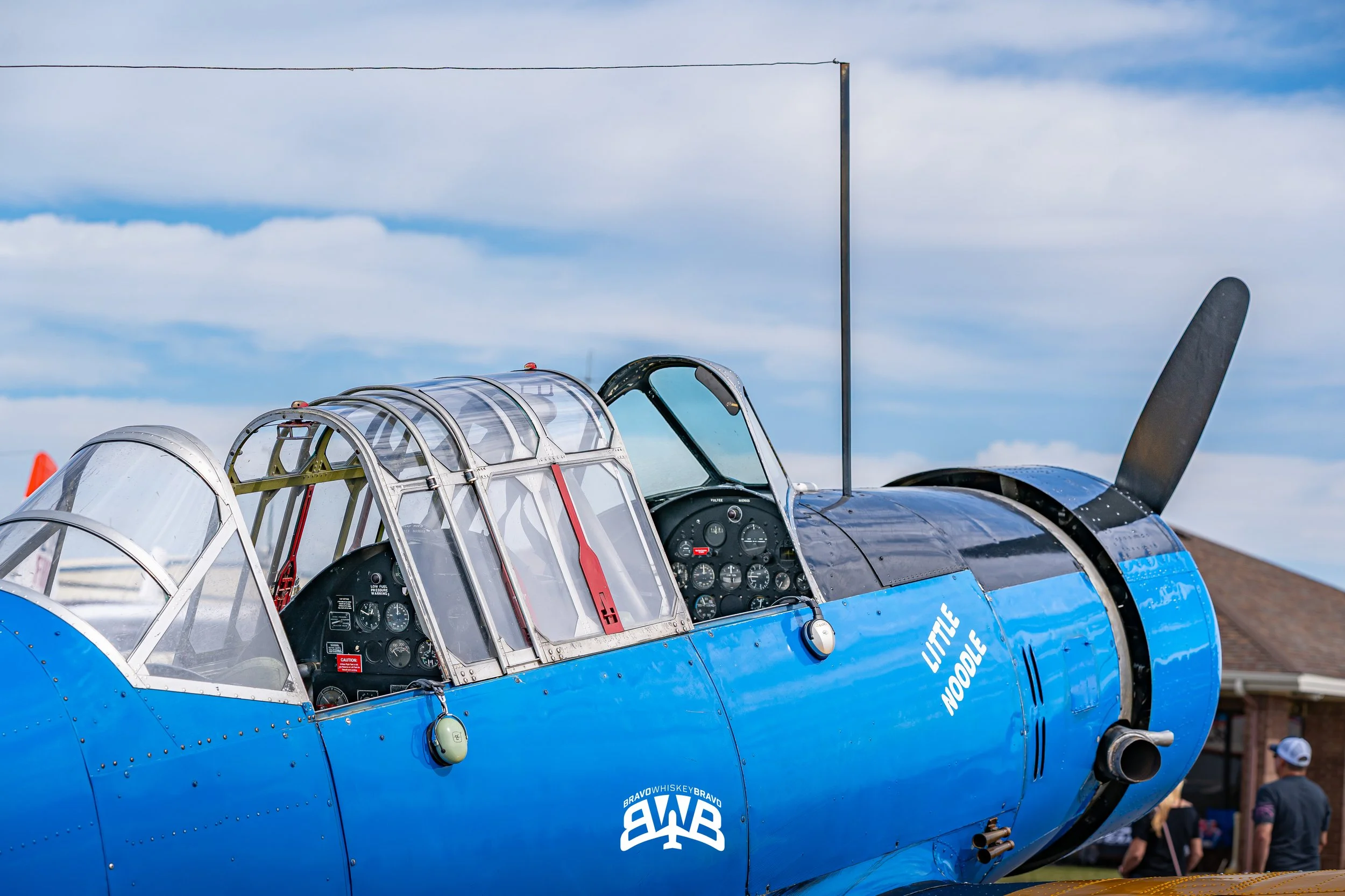 Close-up of a vintage aircraft with a blue body and a cockpit with multiple windows, exposed instruments, and an propeller at the front. The aircraft has the words "LITE NOOLE" and a logo with the text "BRAVO WHISKEY BRAVO" on the side. The sky is pa
