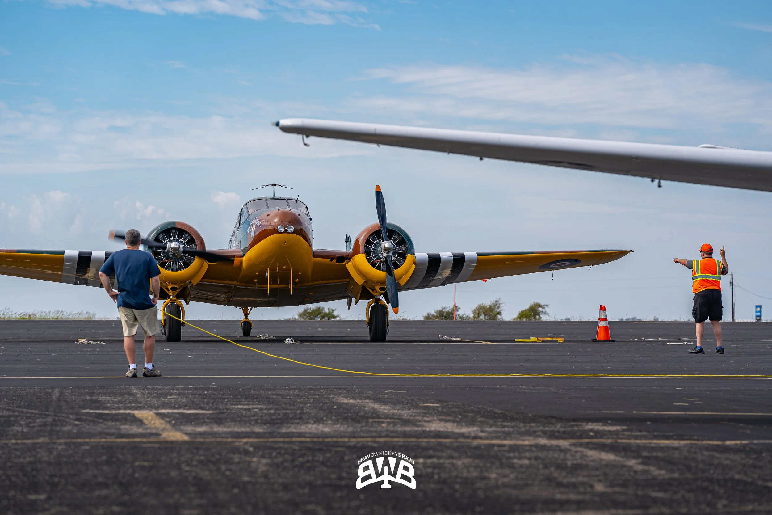 A vintage aircraft with yellow and brown coloring on an airport tarmac, with a man in shorts and a T-shirt on the left and a man in a safety vest and cap on the right directing traffic, with orange cones and a cloudy sky in the background.
