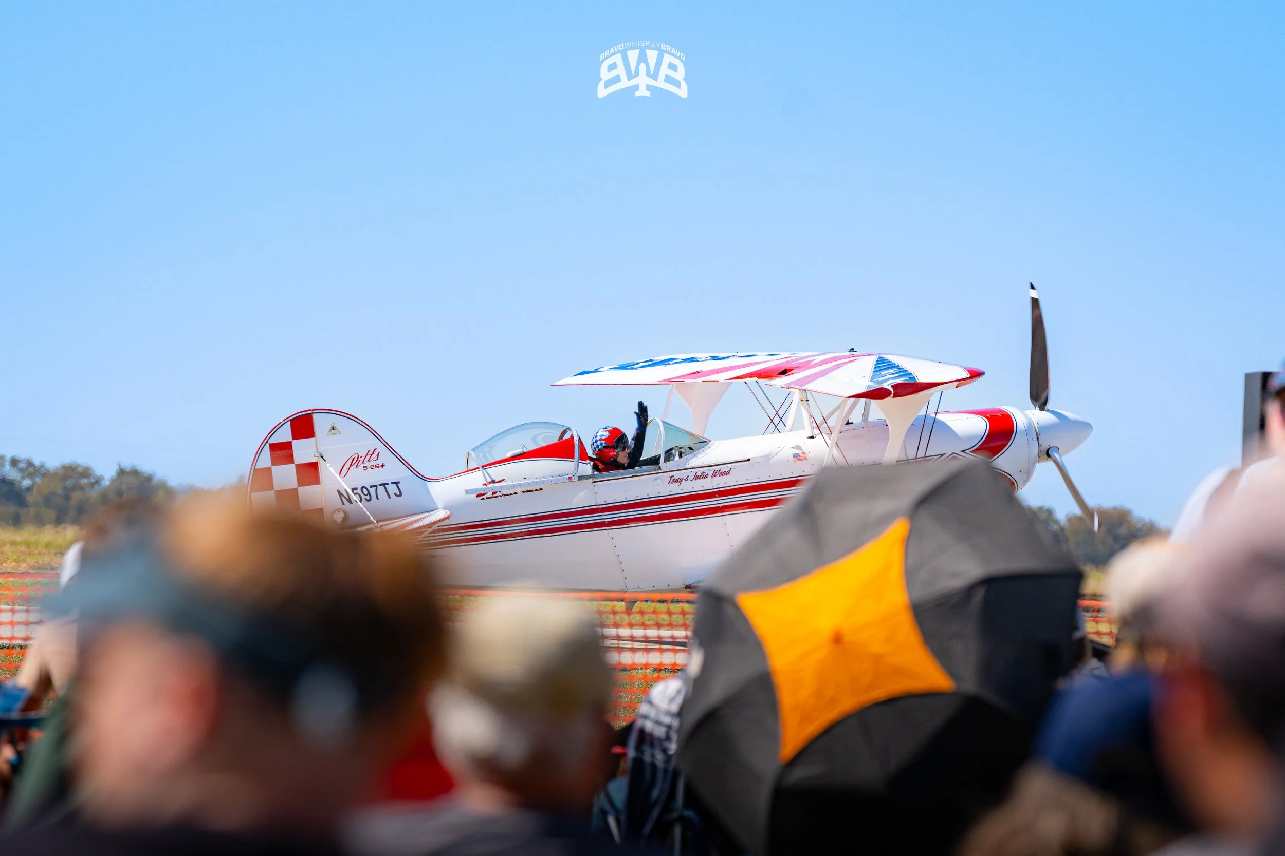 Small white plane with red accents on runway, crowd gathered with some holding umbrellas, pilot waving from open cockpit.