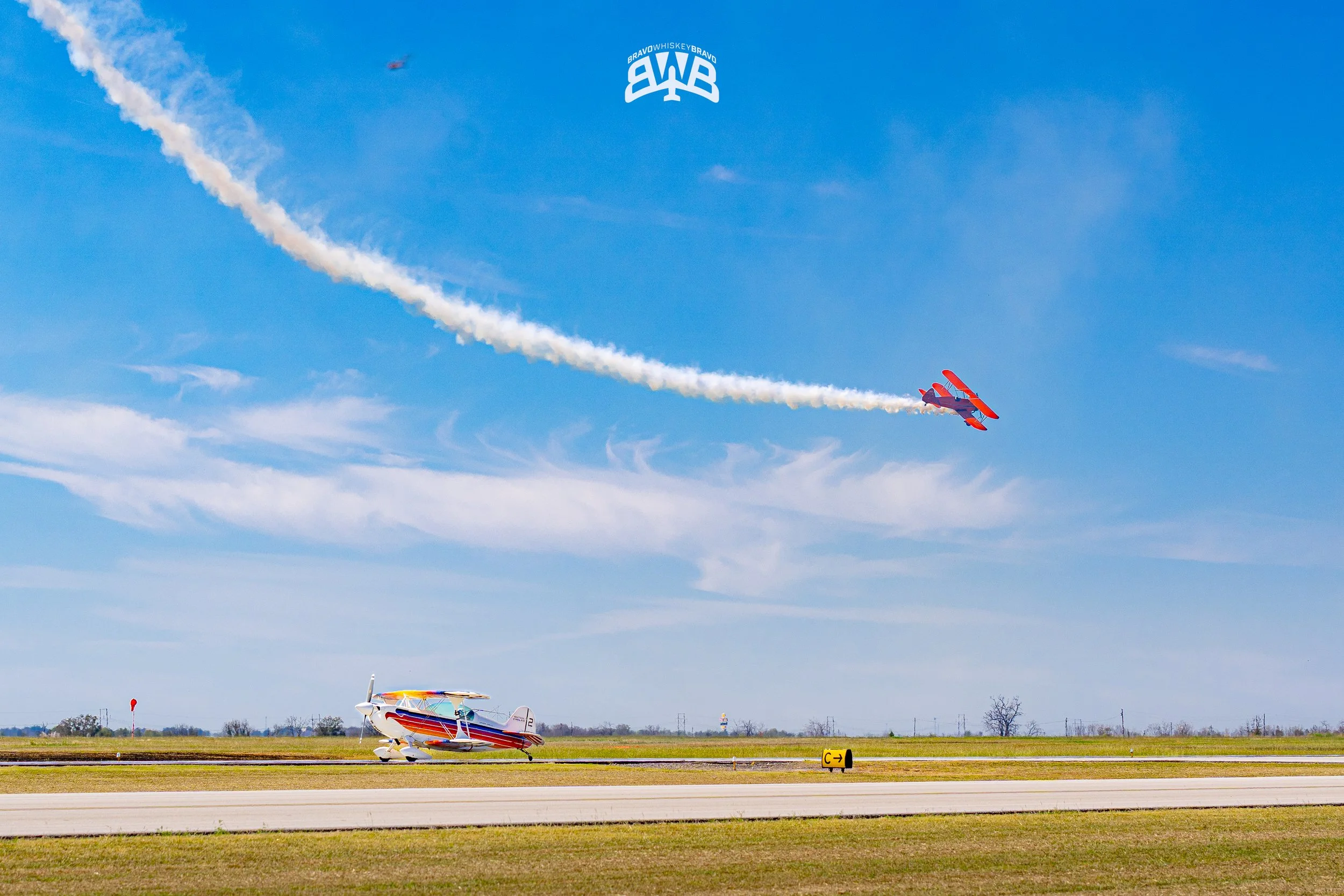 Two colorful aerobatic planes perform a stunt on an airfield, one flying in the sky emitting a white smoke trail, the other resting on the ground under a clear blue sky.