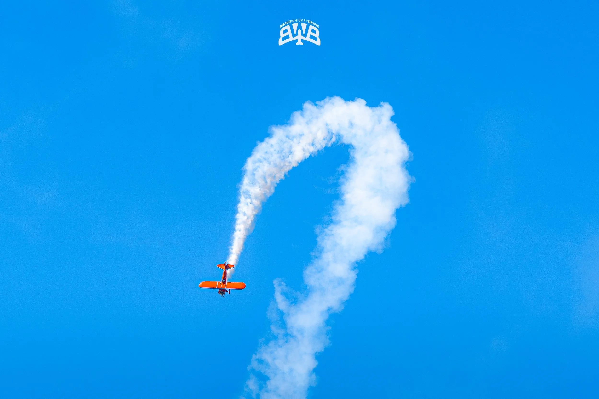 Orange stunt plane flying upside down with white smoke trail forming a question mark against a blue sky, with a blue and white logo at the top.