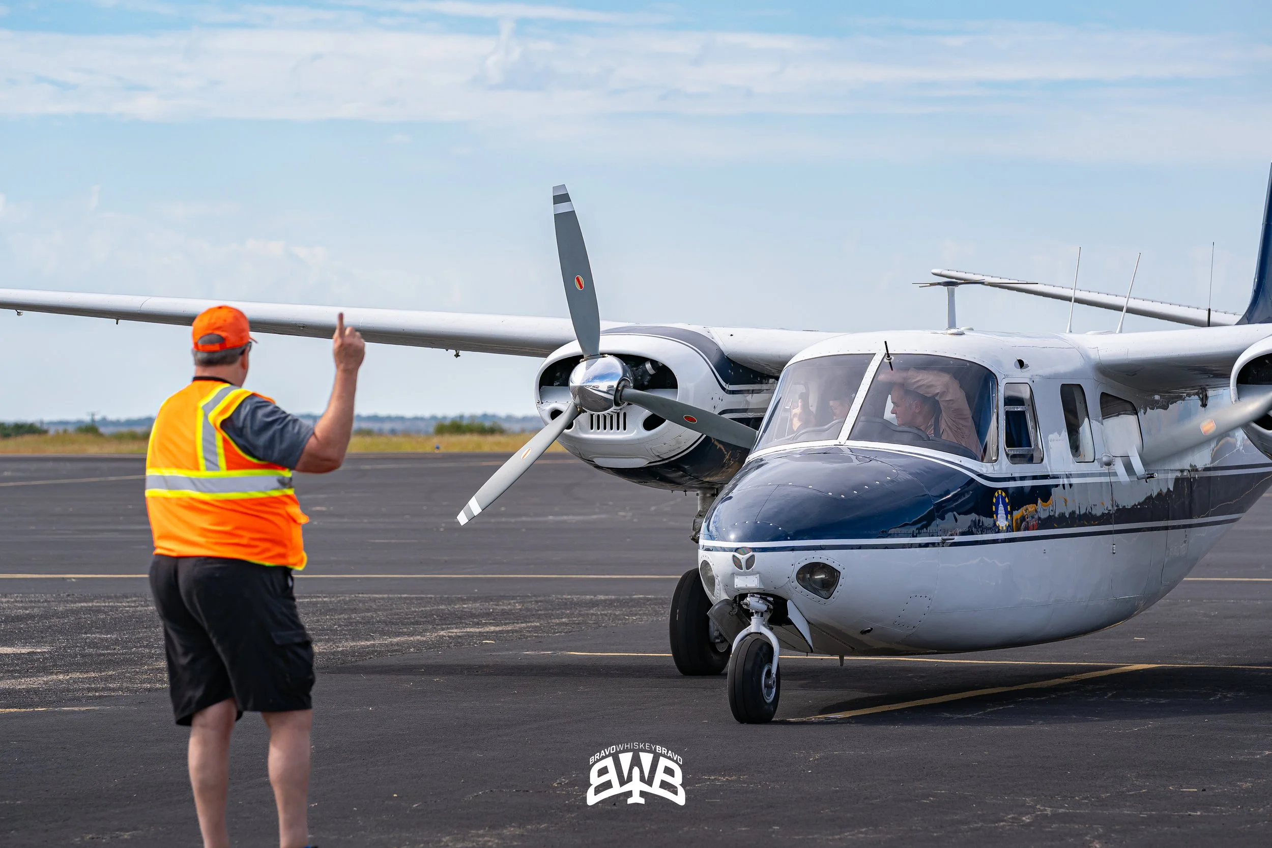 A man in a high visibility orange vest and hat is gesturing with one finger towards a small airplane on the runway, with the plane's pilot visible inside.