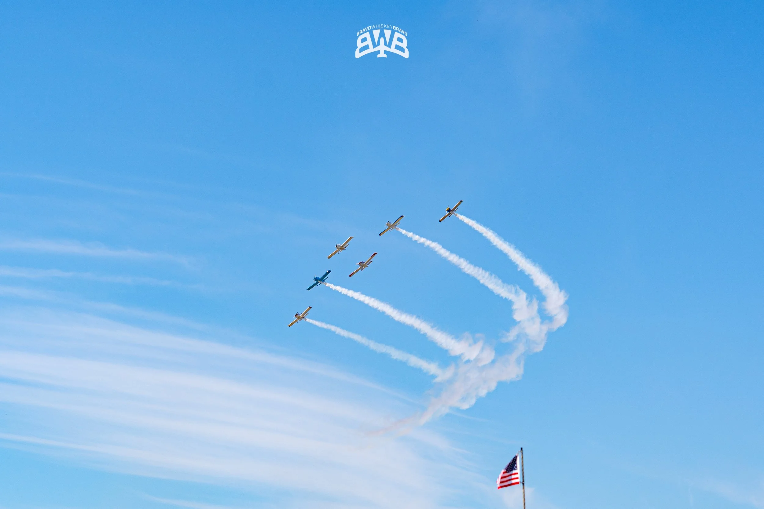 Five airplanes performing a maneuver in the sky with white smoke trails, the American flag flying at the bottom right corner.