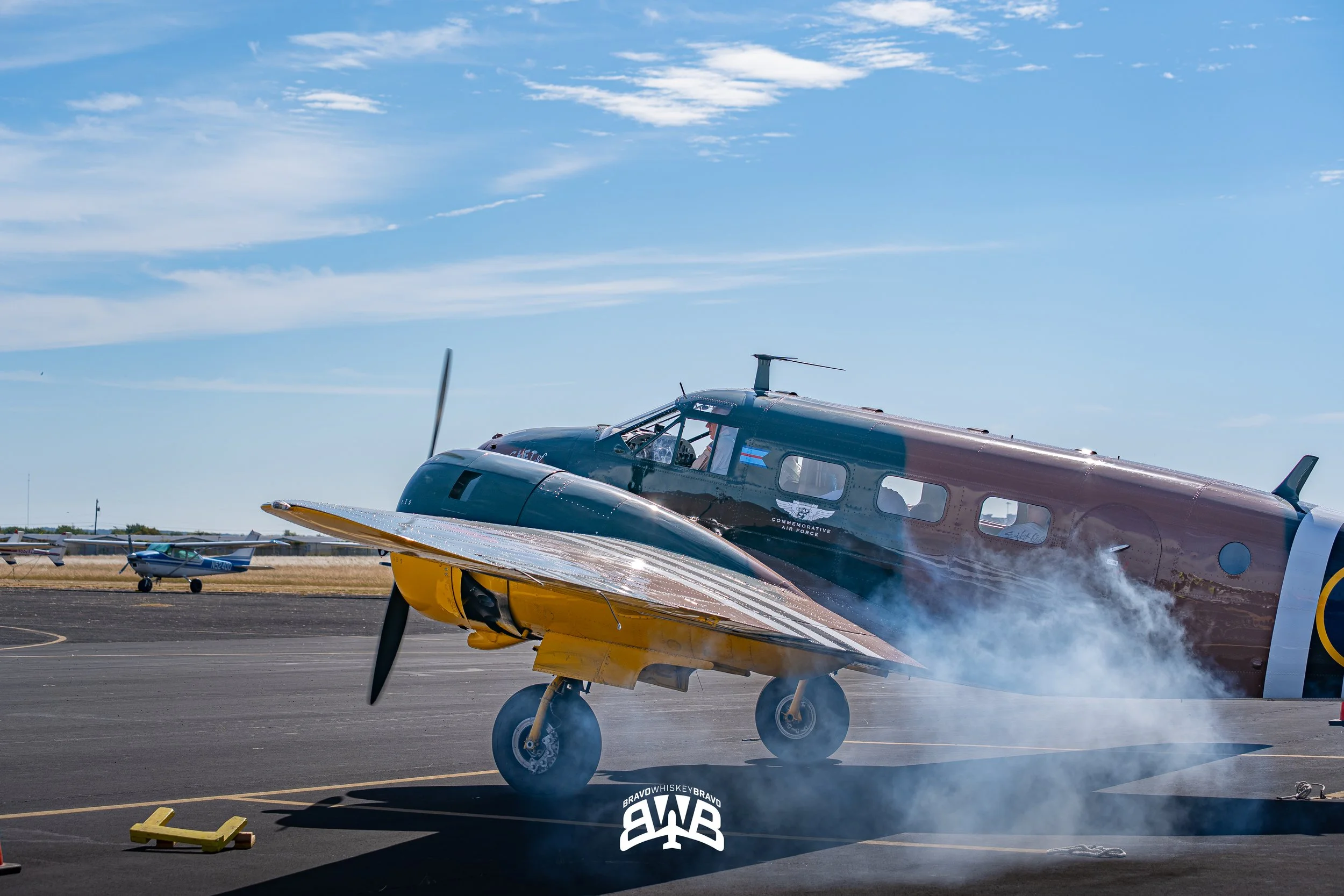Vintage aircraft performing a burnout on an airstrip with smoke, with a small plane in the background under a blue sky.