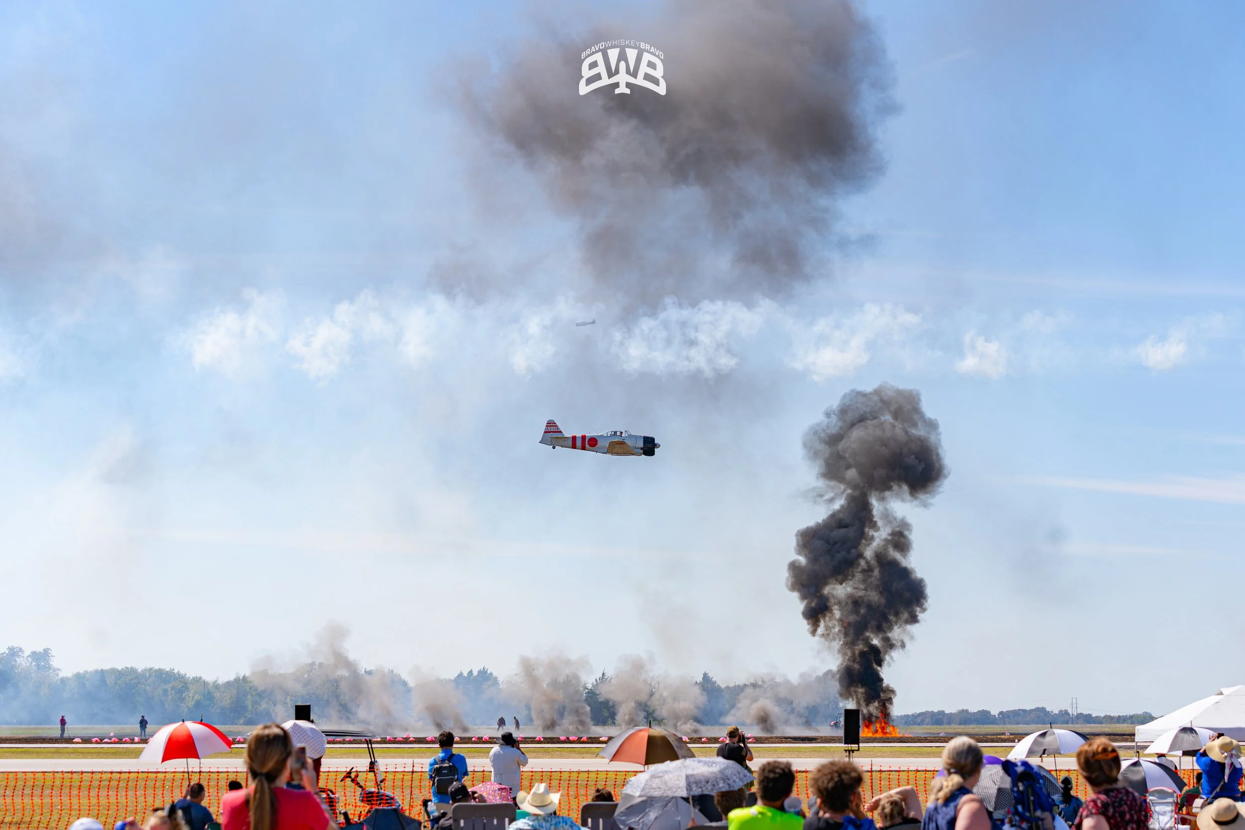 An airshow scene with a crowd of spectators seated on the ground under umbrellas, watching smoke and fire on the runway. Two vintage planes are flying in the sky, one releasing smoke while the other is nearby. Large clouds of black smoke and flames a