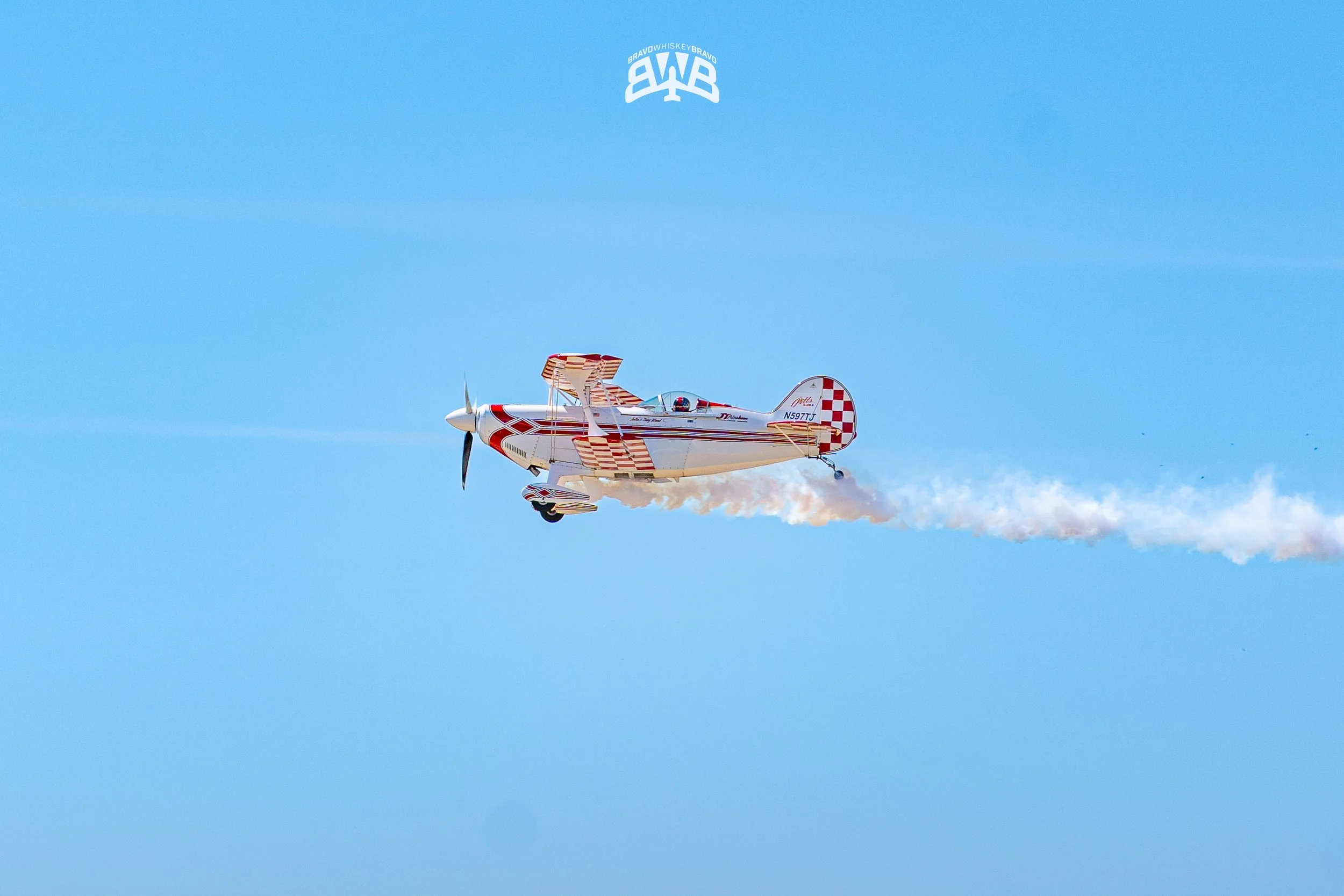 A small red and white airplane flying in a clear blue sky, releasing white smoke.