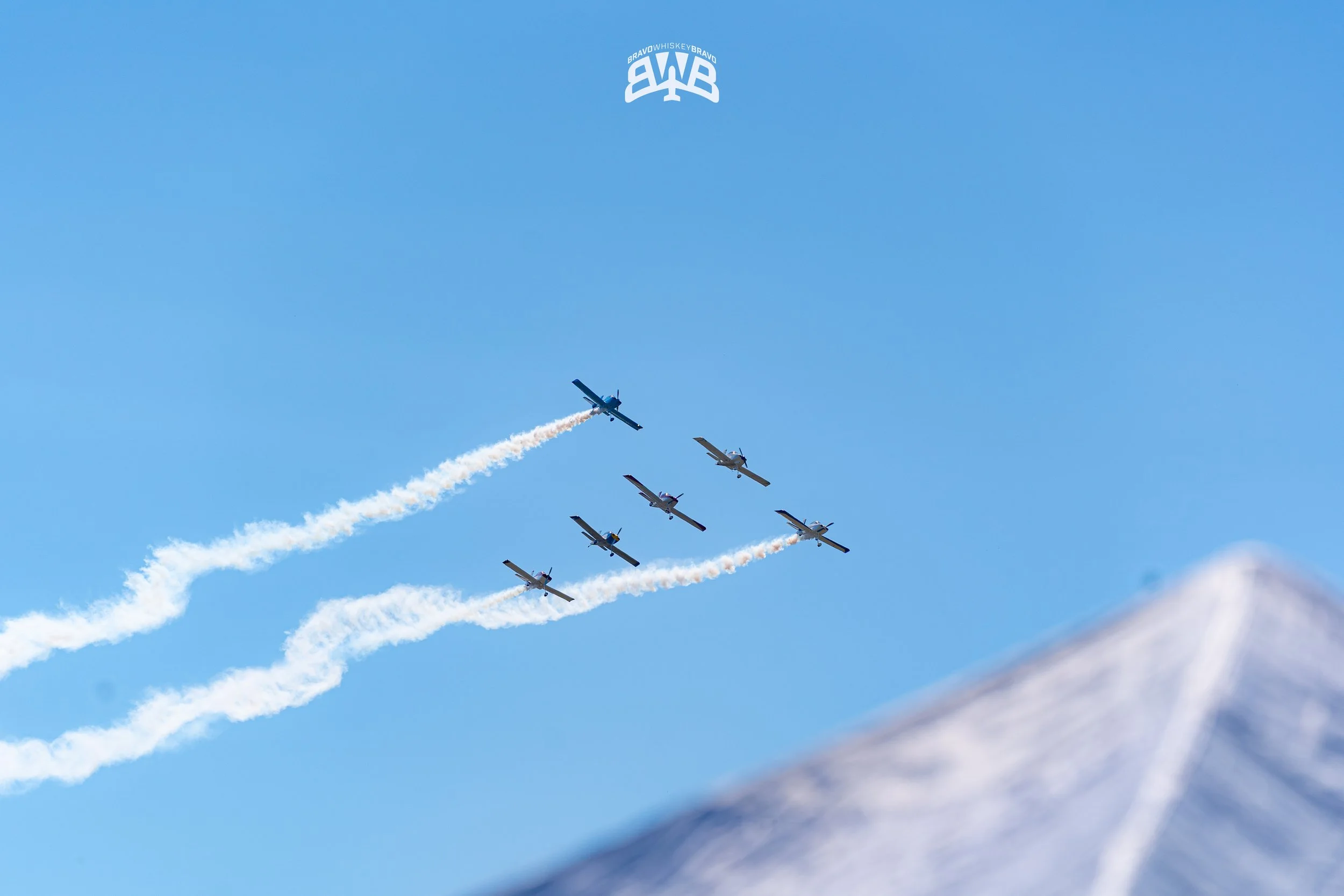 Six airplanes performing an aerial stunt, flying in formation with trails of white smoke against a clear blue sky, with a snowy mountain peak visible in the lower right corner.