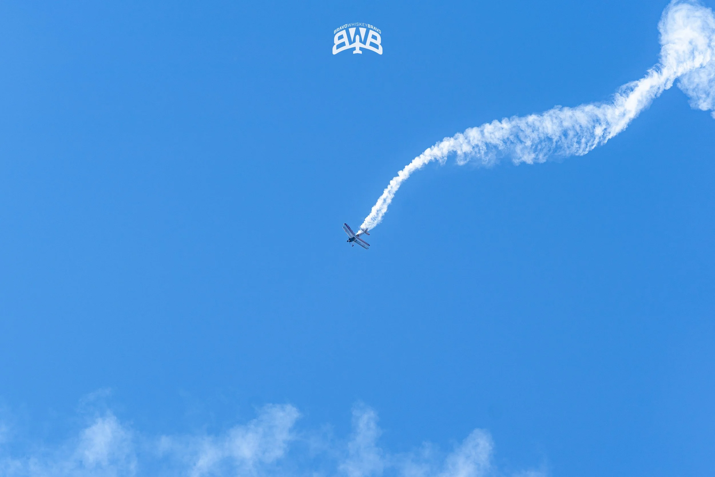 An airplane performing an aerobatic stunt in the sky, leaving behind a trail of white smoke, with a logo or watermark that says BRAVO WHISKEY BRAVO at the top.
