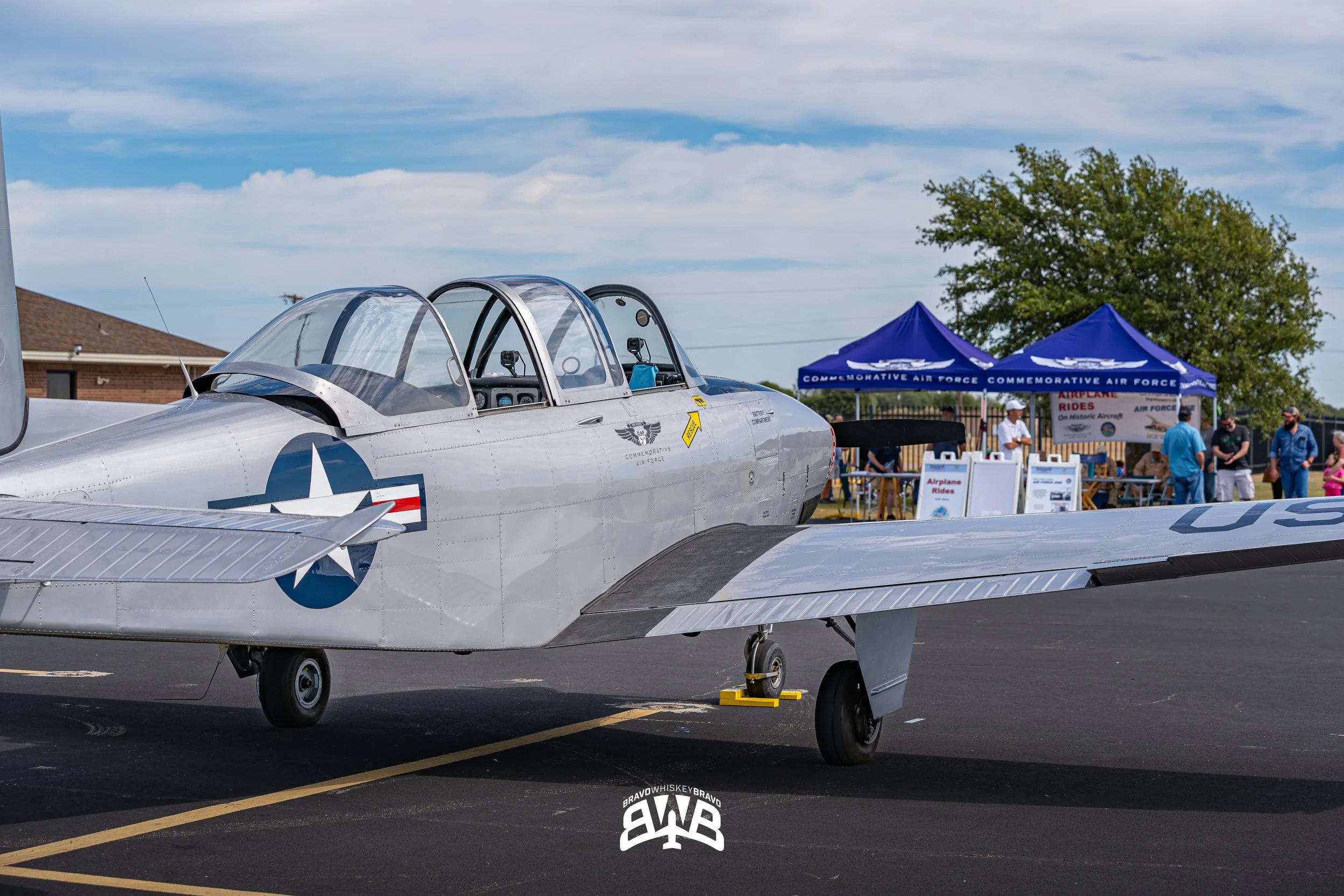 A vintage silver military aircraft on display at an air show, with a blue tent and people in the background.