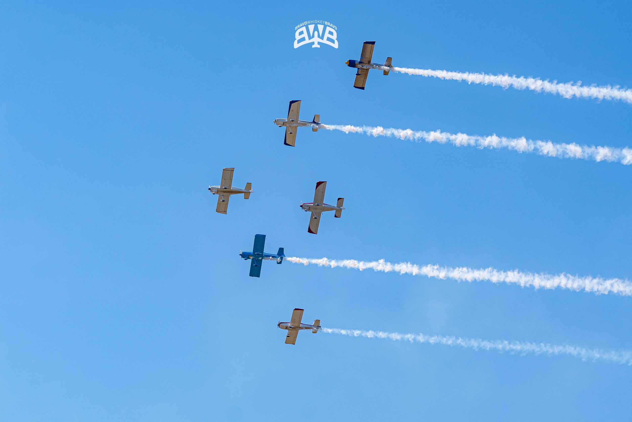 Six small airplanes flying in a circle formation with smoke trails against a bright blue sky, with a logo above them reading BRAVO WHISKEY BRAVO.