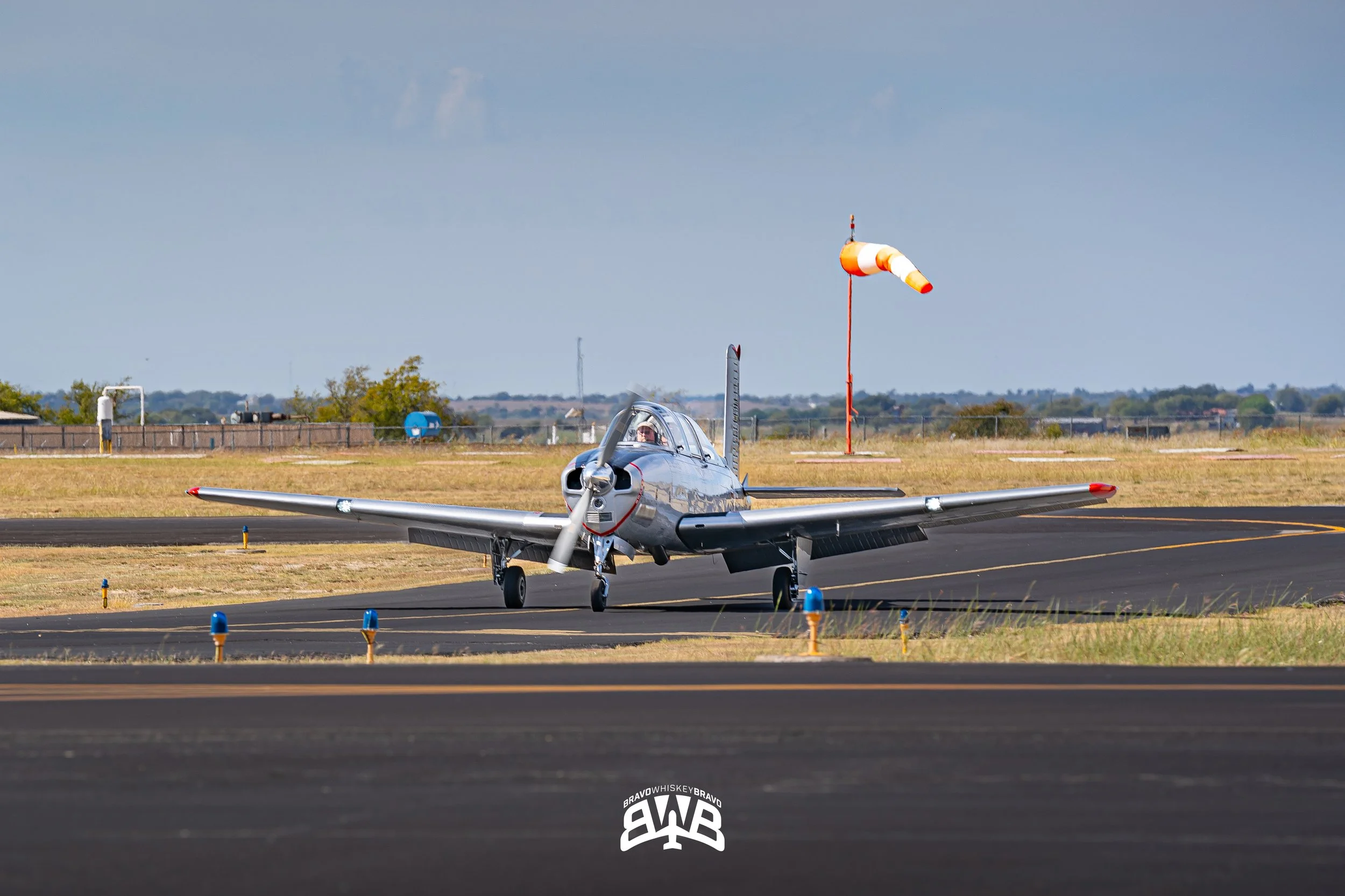 A small, silver propeller airplane on a runway, with a windsock in the background and a partly cloudy sky.