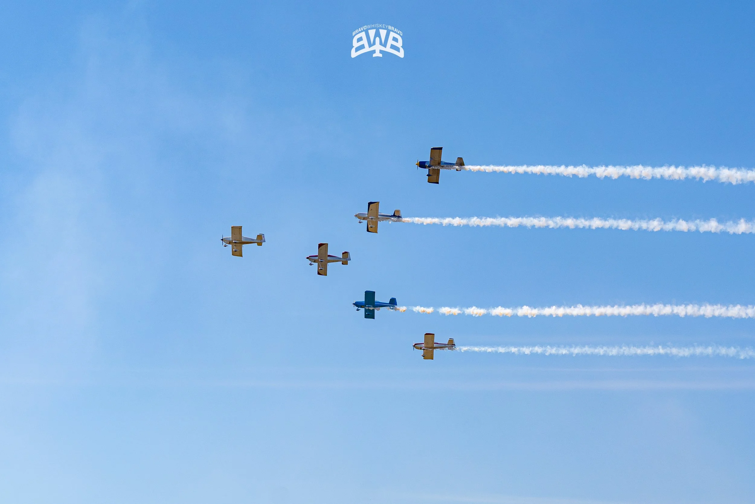 Six small aircraft flying in formation in a clear sky, leaving white smoke trails behind them.