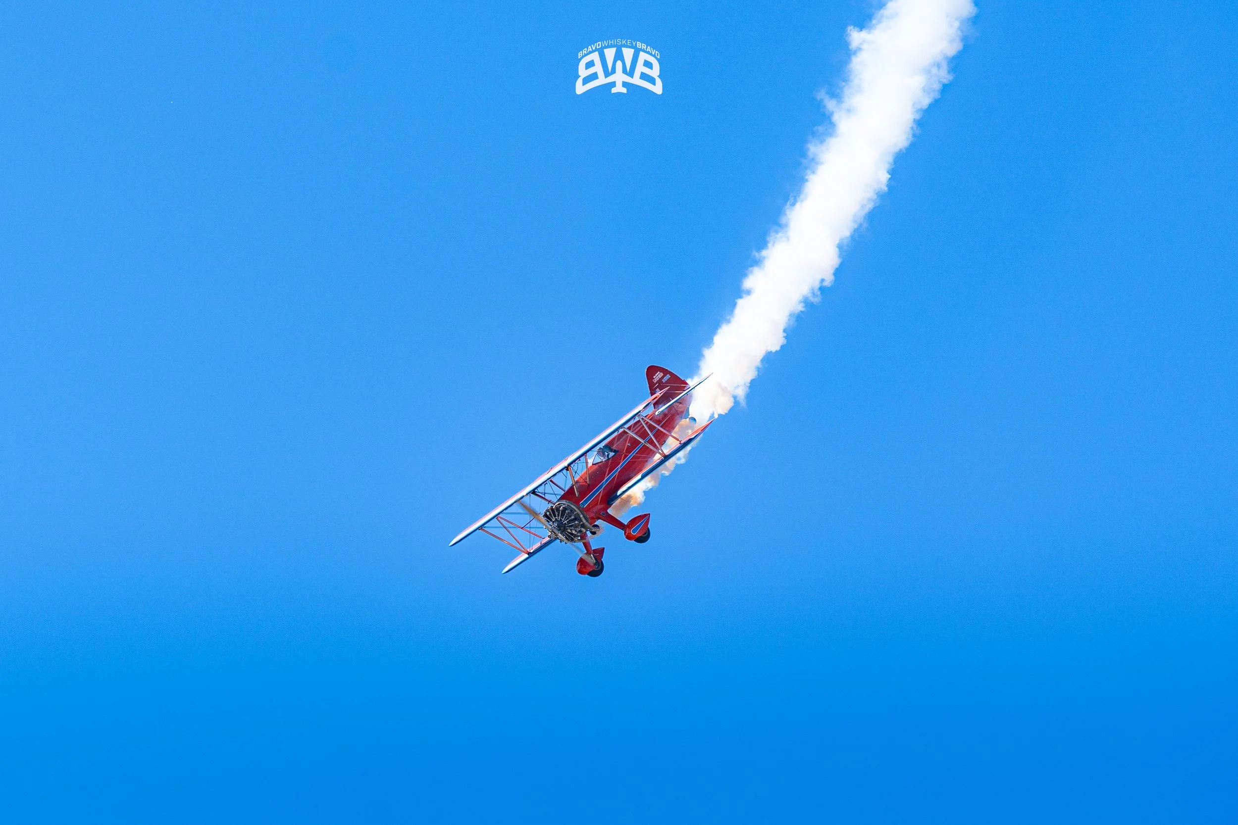 A red biplane performing an aerobatic stunt in the sky, leaving white smoke trail behind on clear blue sky background.