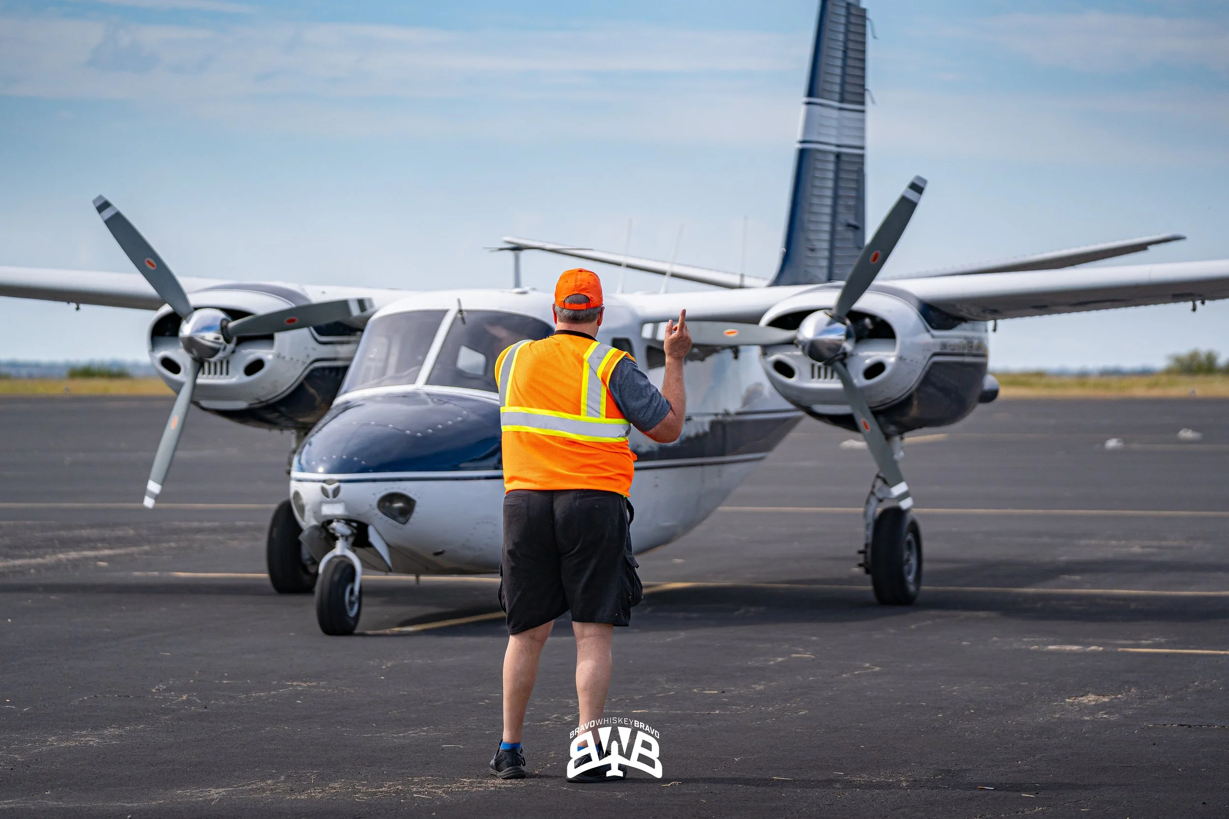 A man in a bright orange safety vest and cap stands on a tarmac, gesturing with one finger, facing a small private airplane with twin engines and propellers, under a partly cloudy sky.