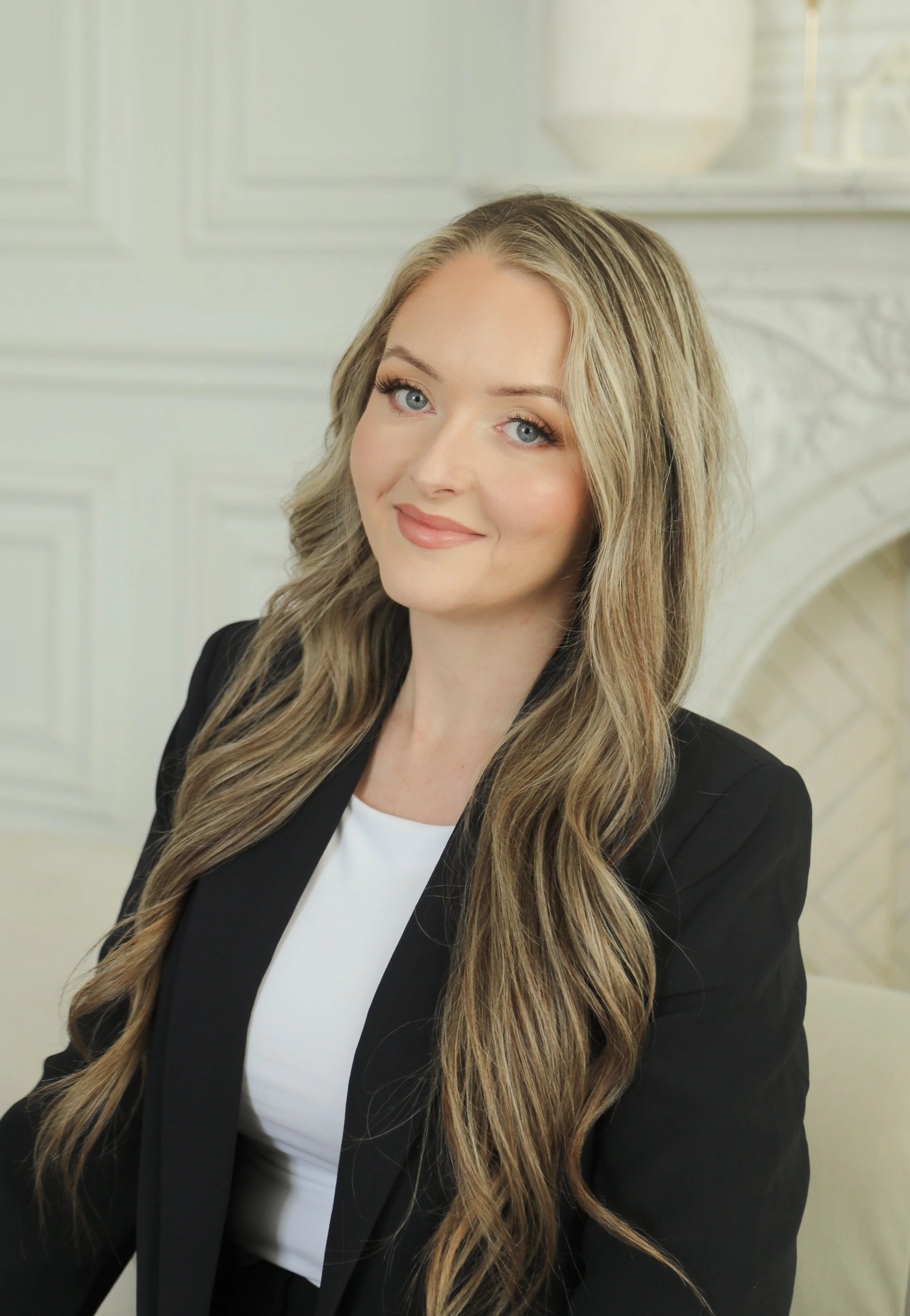 A woman with long, wavy blonde hair, blue eyes, and light makeup, wearing a black blazer and white top, sitting in a bright, elegant room with white paneling and decor.