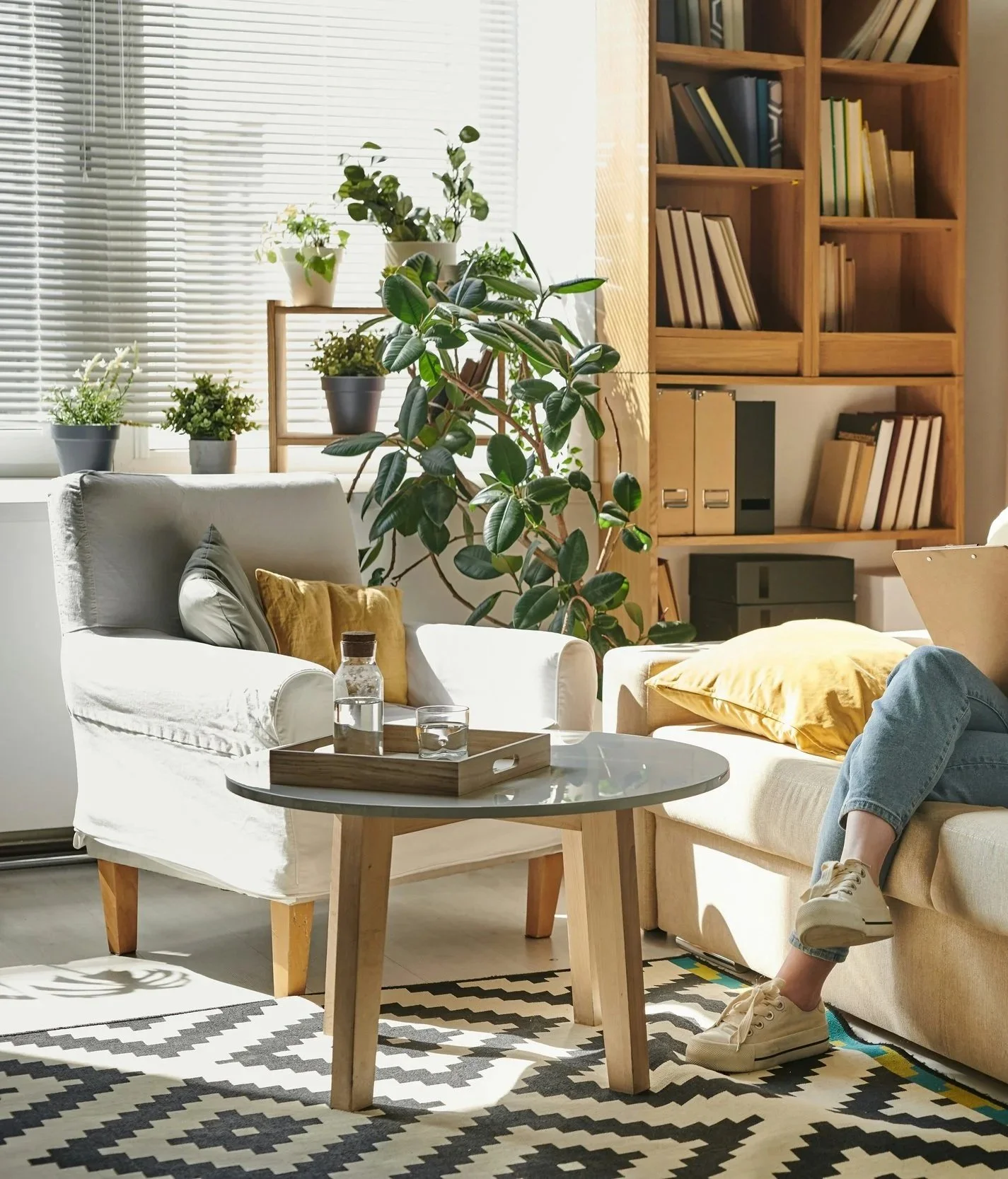 Living room with sunlight, gray sofa, yellow and gray pillows, small round wooden coffee table with water bottle and glass, person sitting on sofa wearing jeans and sneakers, plants on window sill, bookshelf with books and binders.