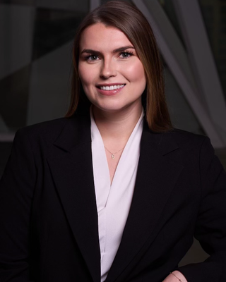 Professional woman with long brown hair wearing a black blazer and white shirt, smiling outdoors.