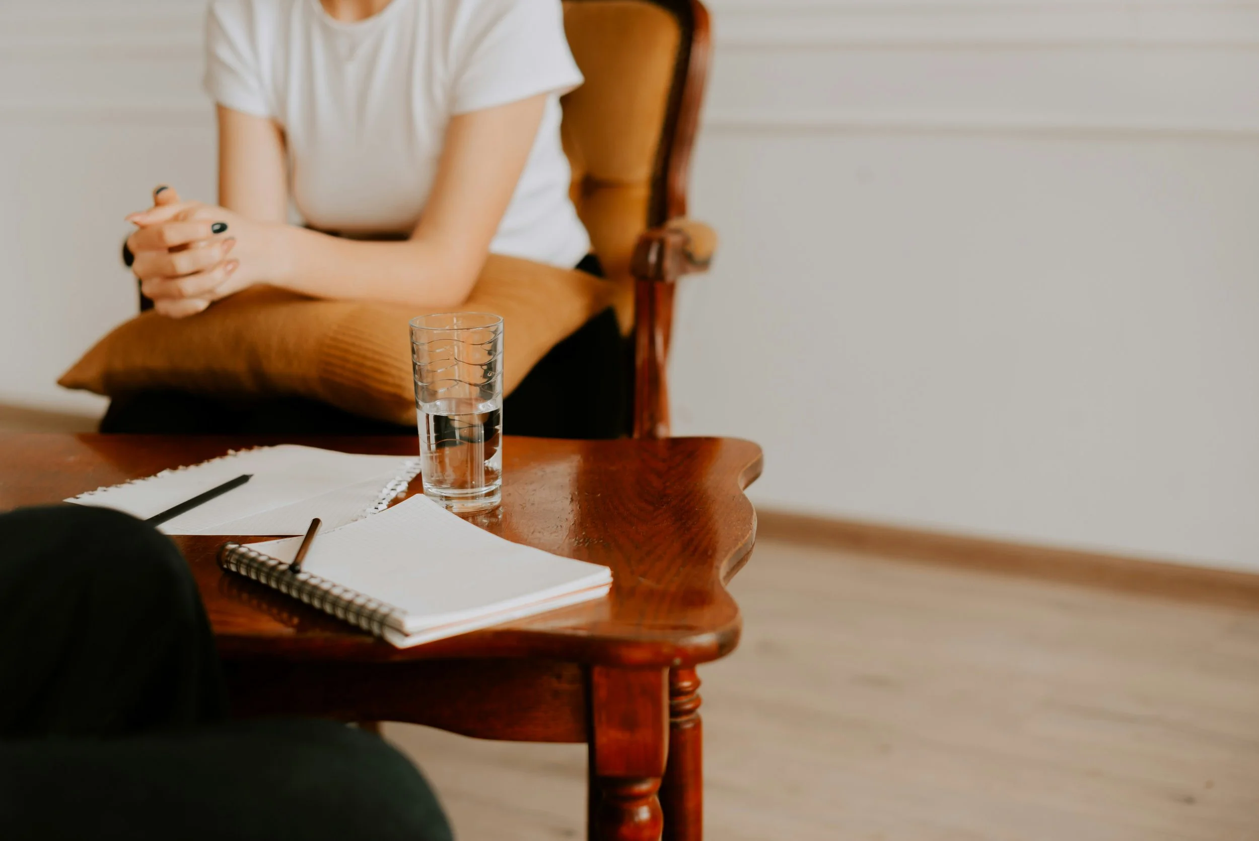 A woman sitting on a chair with her hands clasped, a wooden table with a glass of water, notebooks, and pens, and a person sitting nearby in a room with white walls and Hardwood floors.