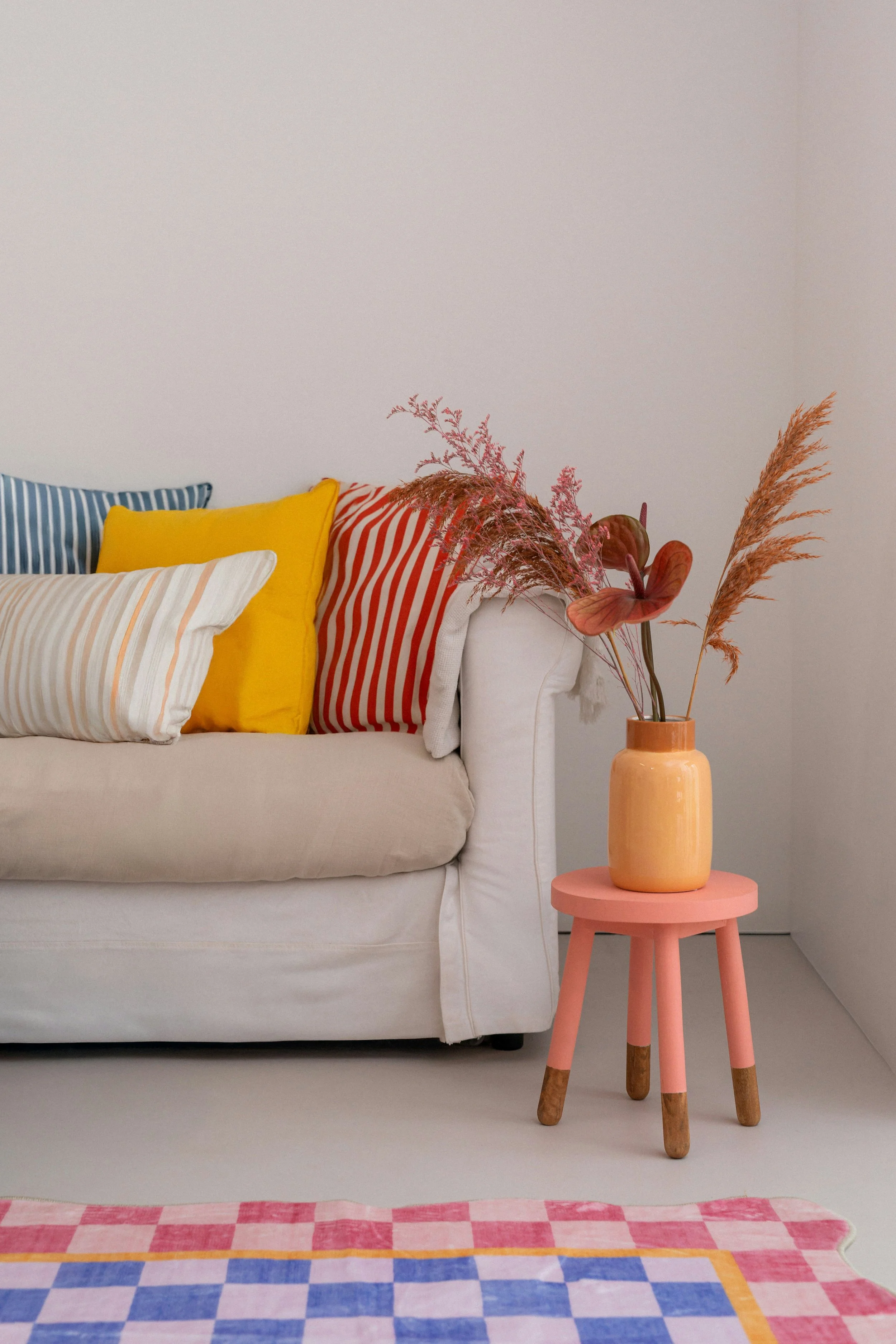 A cozy living room corner with a white sofa decorated with colorful pillows in stripes and solid colors, a pink side table with a yellow vase holding dried ornamental grasses, against a plain white wall, and a colorful checkered rug on the floor.