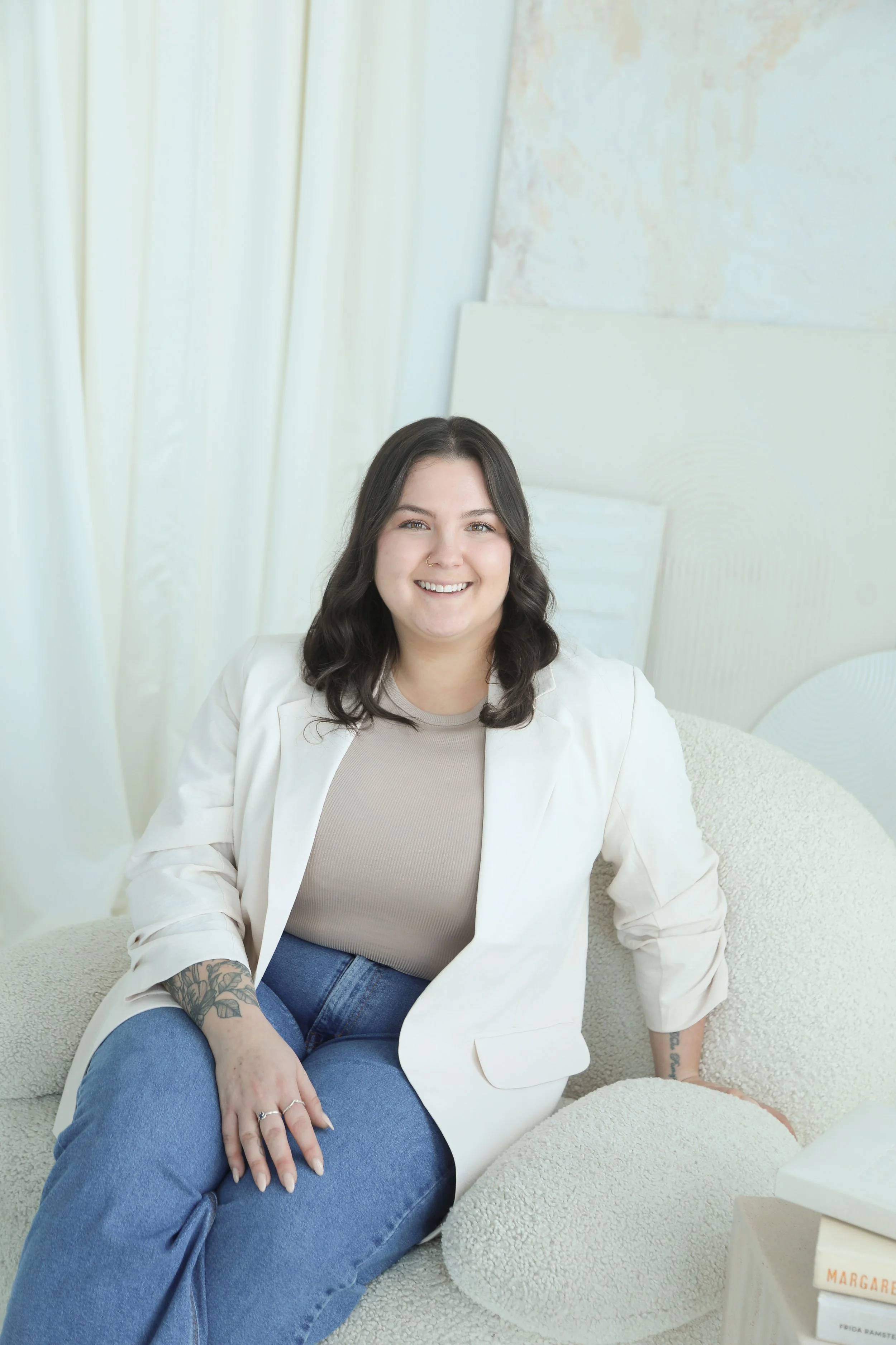 A woman with dark hair wearing a light-colored blazer and jeans sitting on a cream textured sofa, smiling in a bright, modern room.