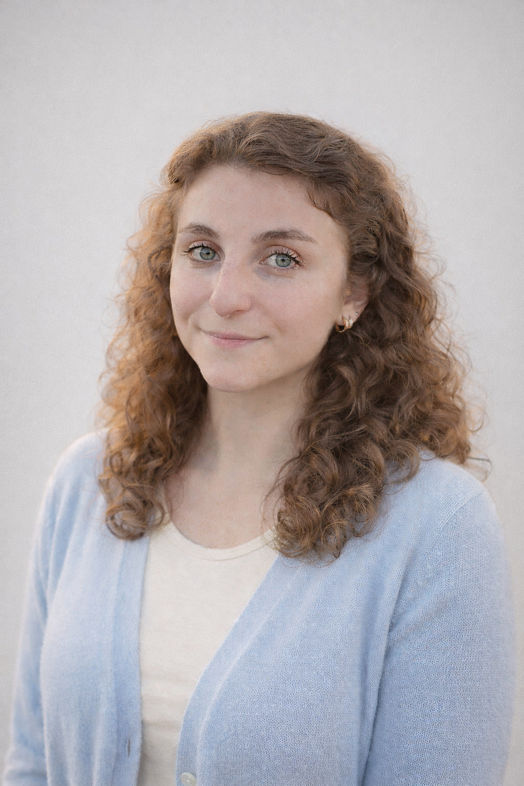A young woman with curly red hair and blue eyes, smiling softly, wearing a light blue cardigan and a cream-colored top, standing against a plain light background.
