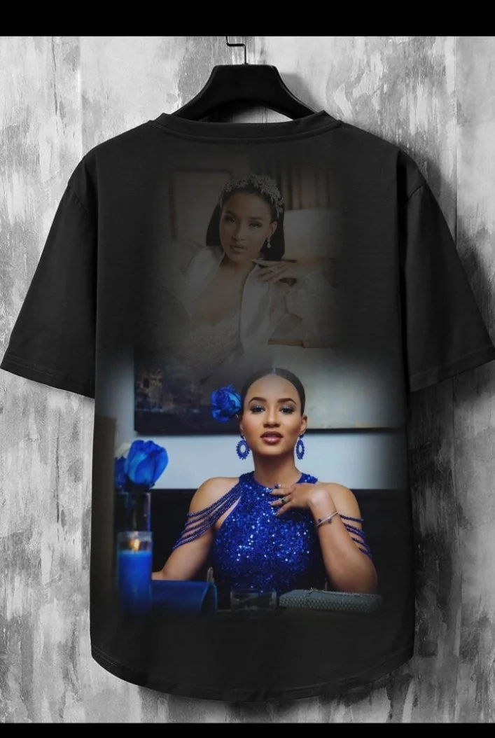 T-shirt noir avec une image de deux femmes, une en haut avec une expression sérieuse, et une en bas portant une robe bleue scintillante, assise à une table avec des fleurs bleues.
