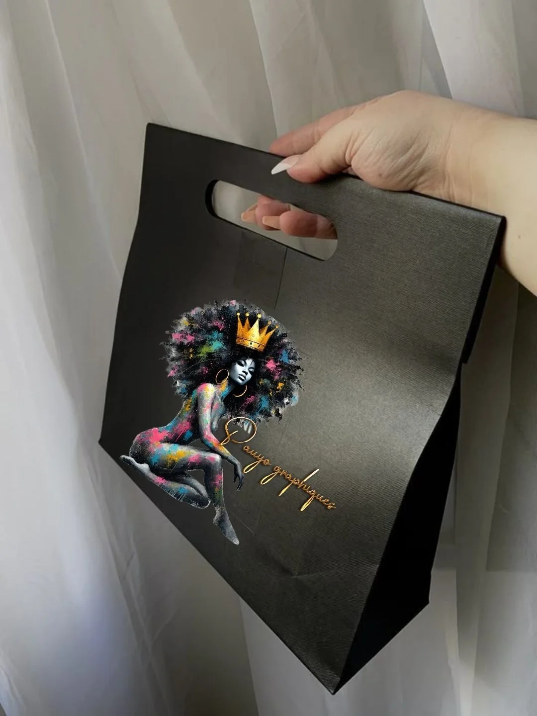 Sac noir avec une illustration d'une femme avec une coiffure afro colorée, portant une couronne et des accessoires dorés, accompagnée du texte "Dauya graphismes" en or.