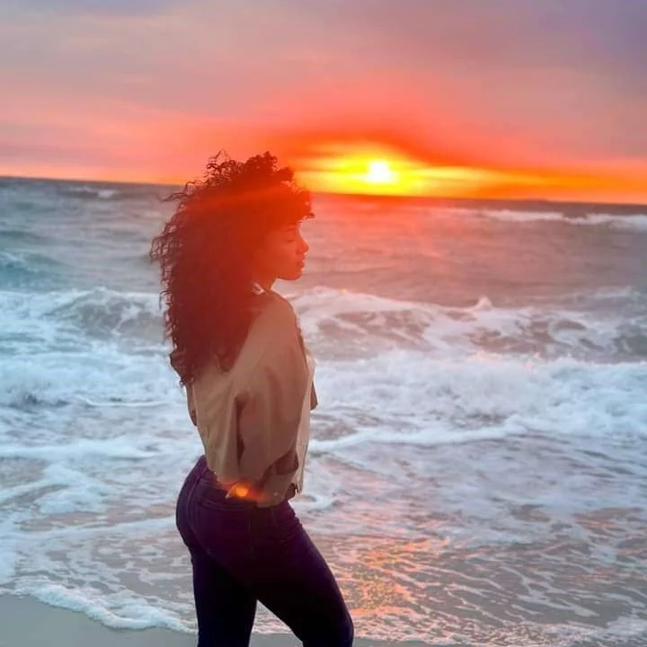 Une femme avec des cheveux bouclés regarde la mer au coucher du soleil, sur une plage.
