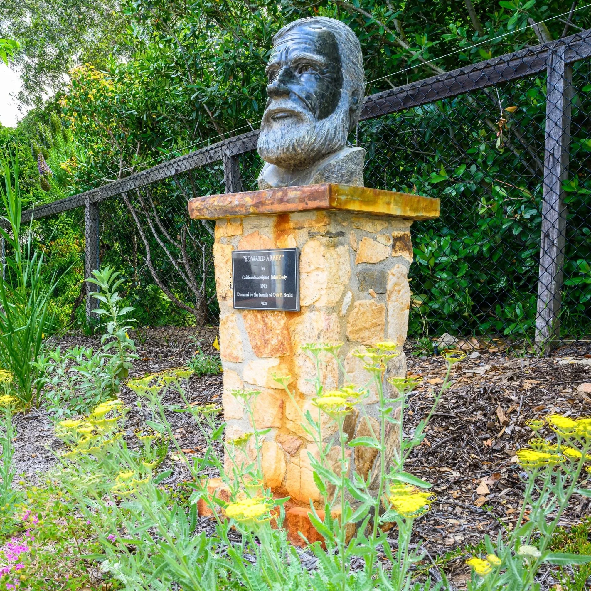 Bronze bust of a man with a beard on a stone pedestal, surrounded by plants and flowers, with a black fence in the background.