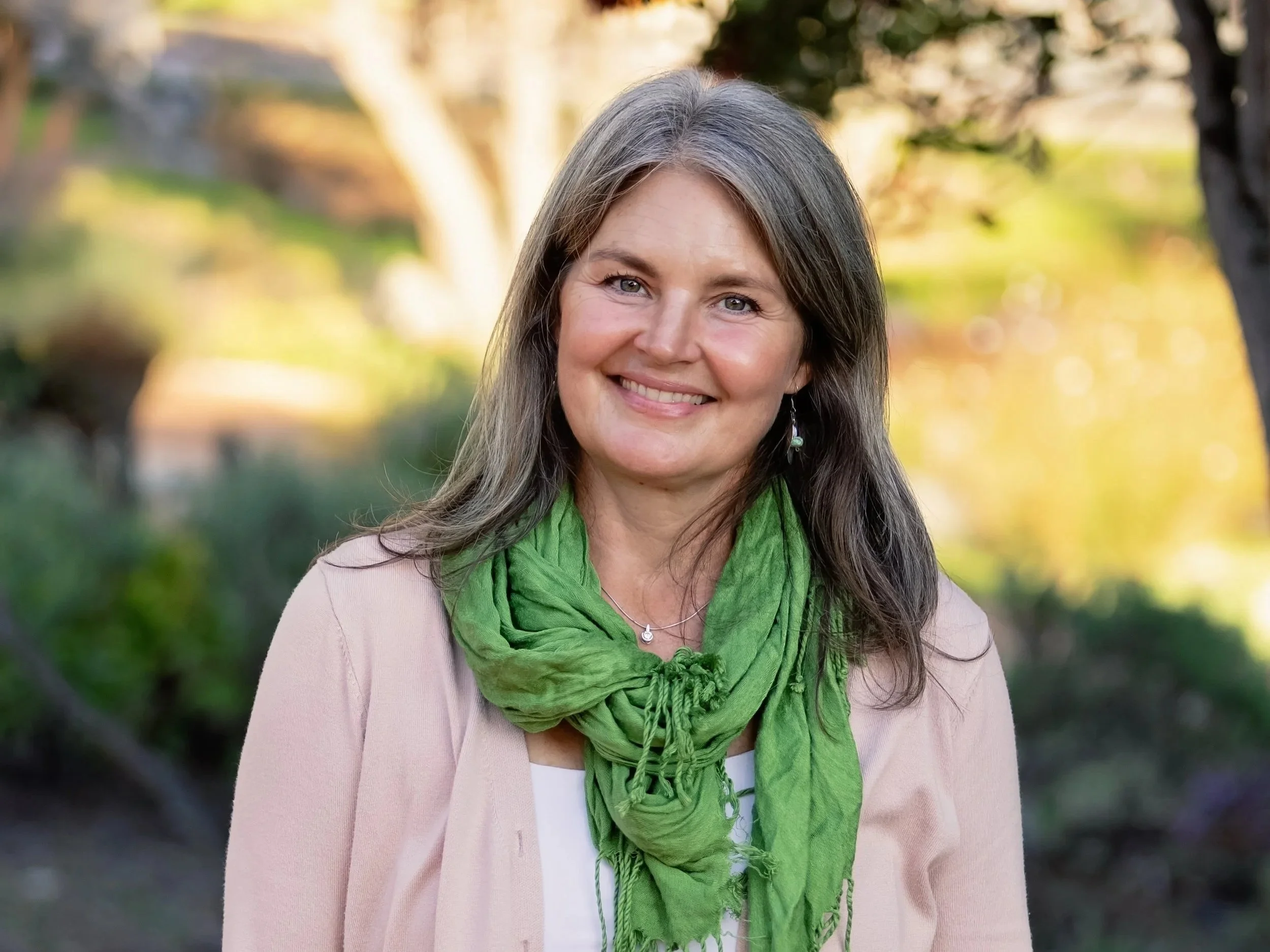 A woman with long gray hair wearing a beige blazer, white top, and green scarf, smiling outdoors with trees and blurred autumn foliage in the background.