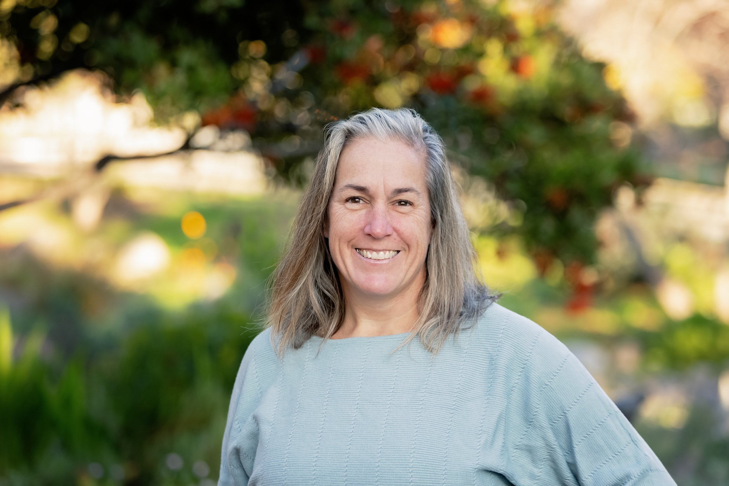 A woman with shoulder-length gray hair smiling outdoors with blurred trees and foliage in the background.