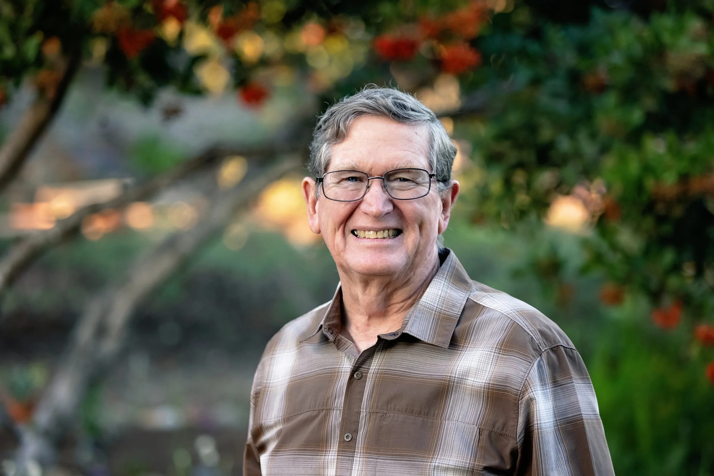 An older man with gray hair and glasses smiling outdoors, wearing a plaid shirt with trees and sunlight in the background.