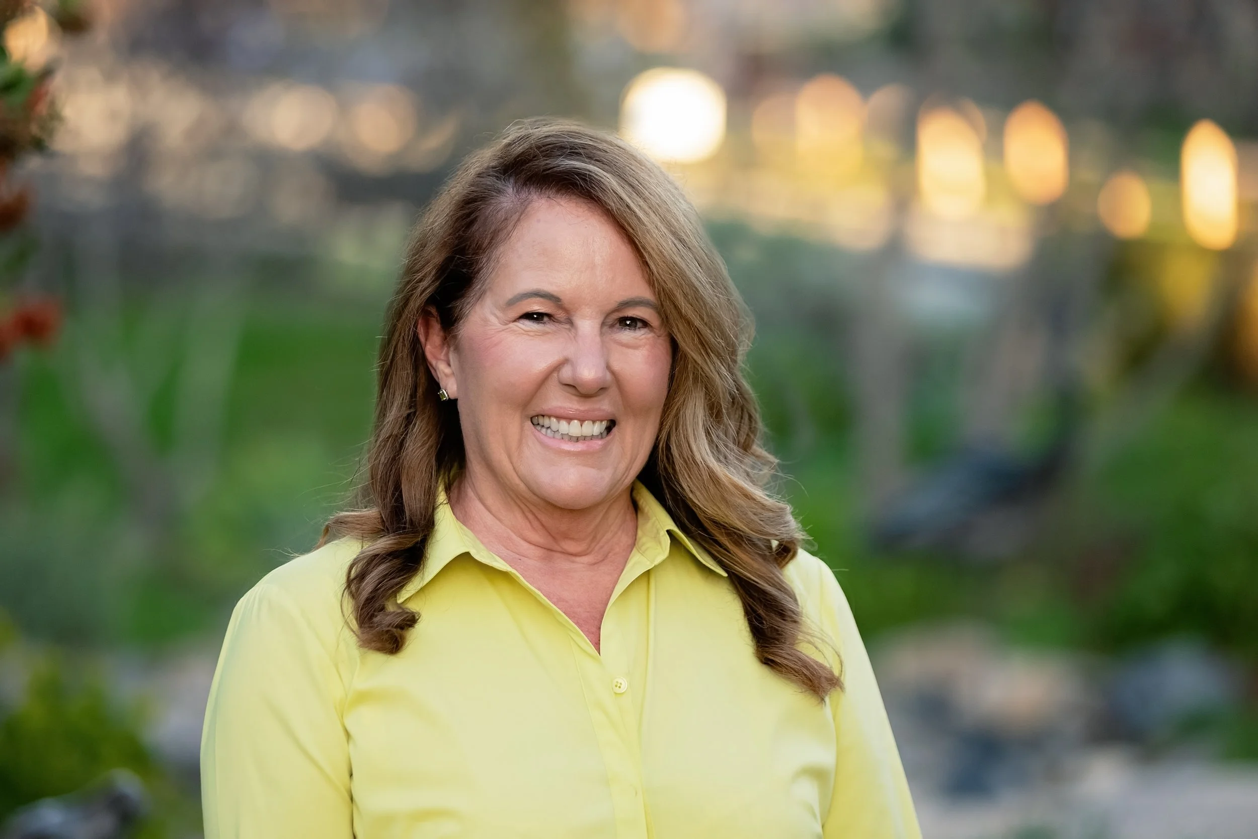 A woman with long, wavy brown hair smiling in an outdoor setting with blurred greenery and bokeh lights in the background, wearing a light yellow button-up shirt.