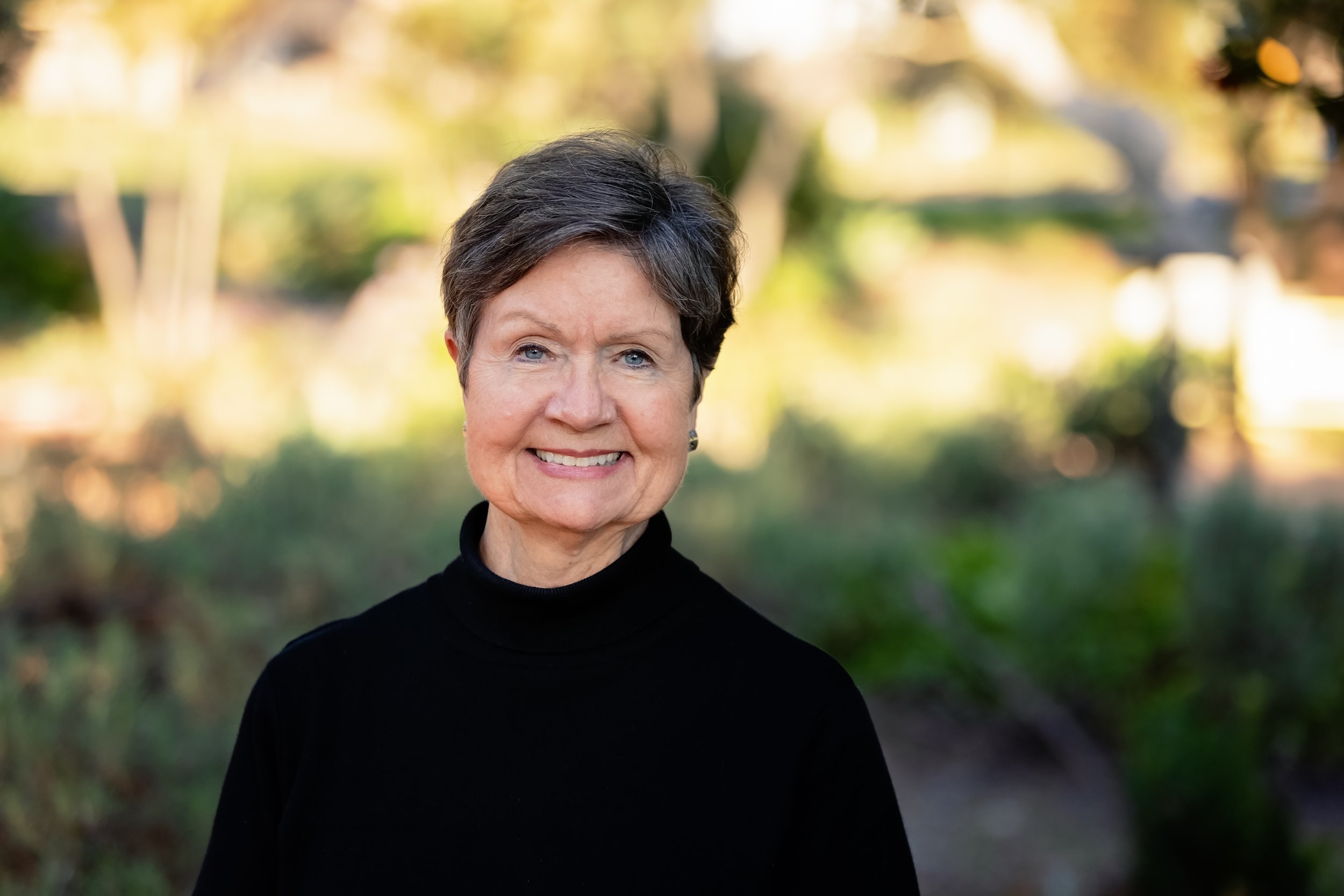 Portrait of an elderly woman with short gray hair, wearing a black turtleneck sweater, smiling outdoors with blurred greenery and trees in the background.