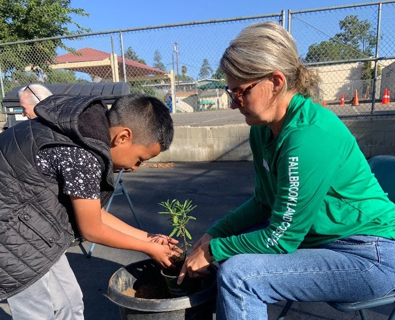 A woman and a boy planting a young tree into a pot outside on a sunny day.