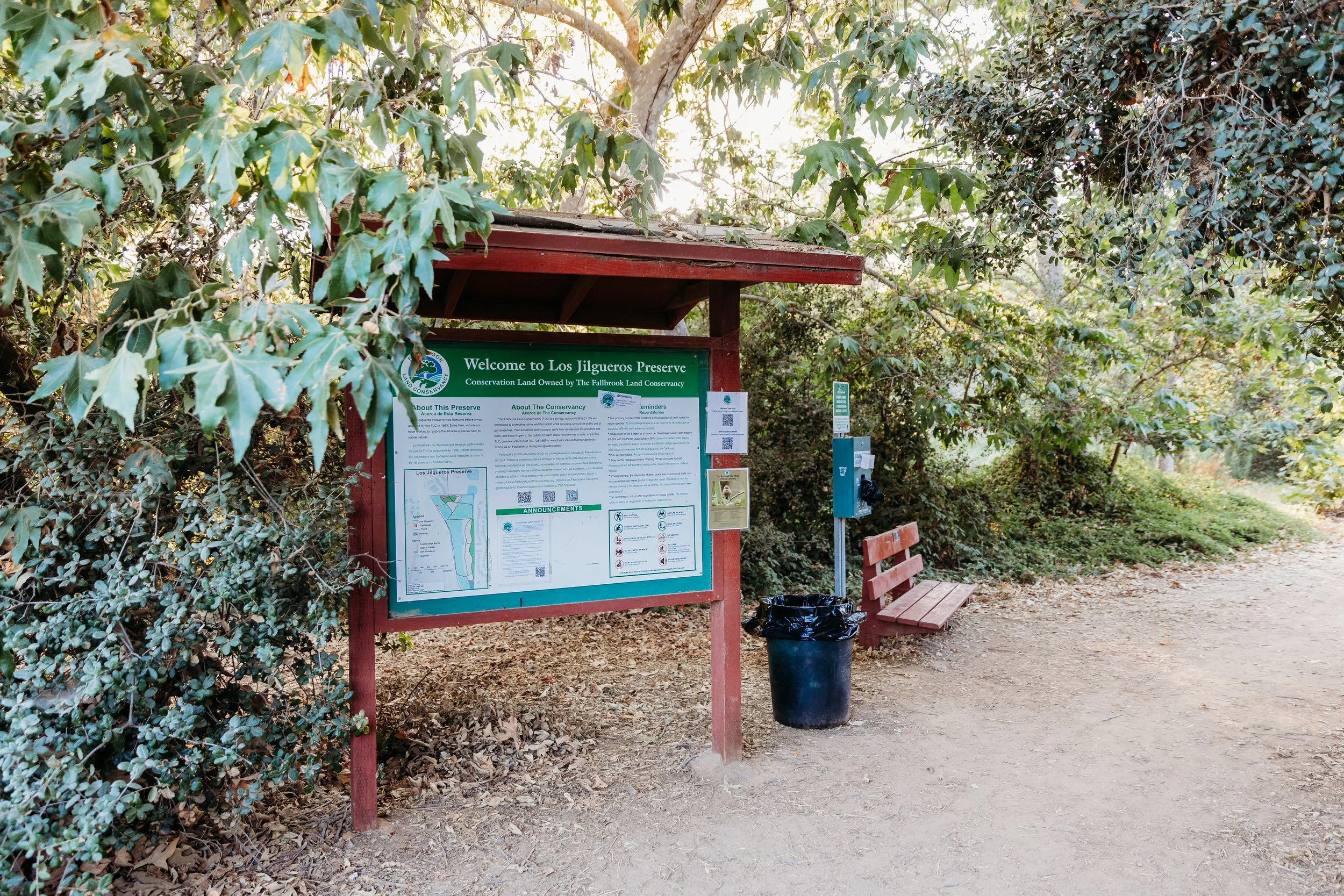 Information board at Los Jilgueros Preserve with a bench, a trash can, and surrounding trees and bushes.