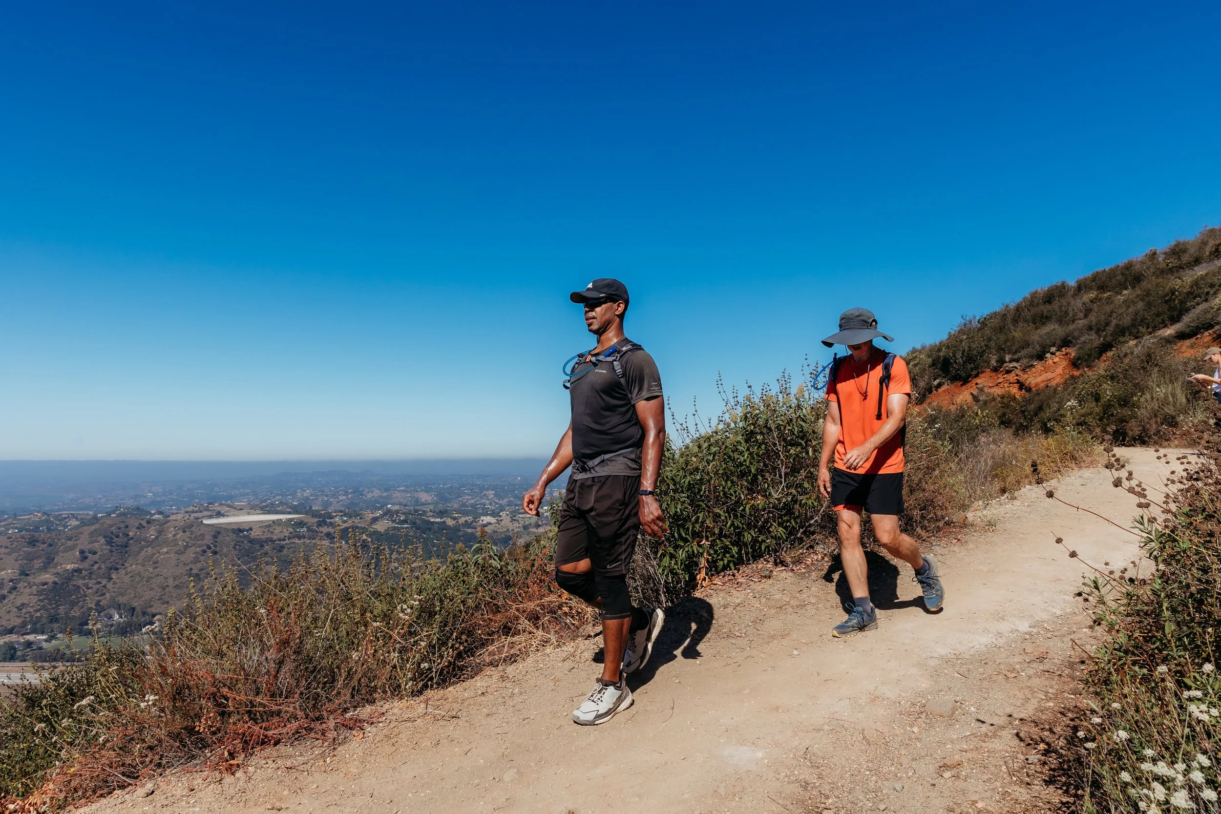 Two men hiking on a dirt trail on Monserate Mountain with a view of a valley and cityscape in the distance under a clear blue sky.