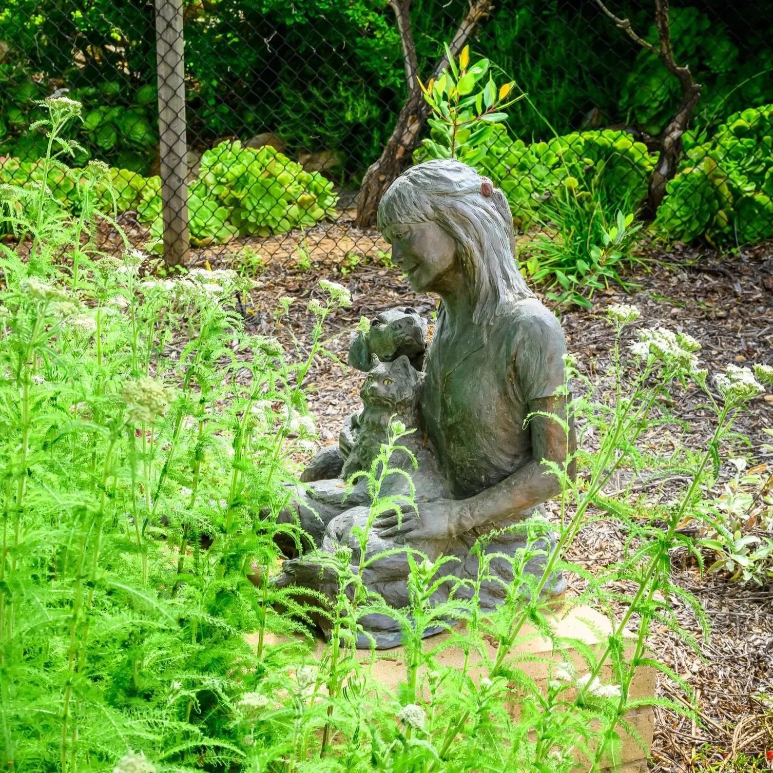 A bronze statue of a girl sitting in a garden, surrounded by green plants, holding a small dog in her lap with other animals nearby.